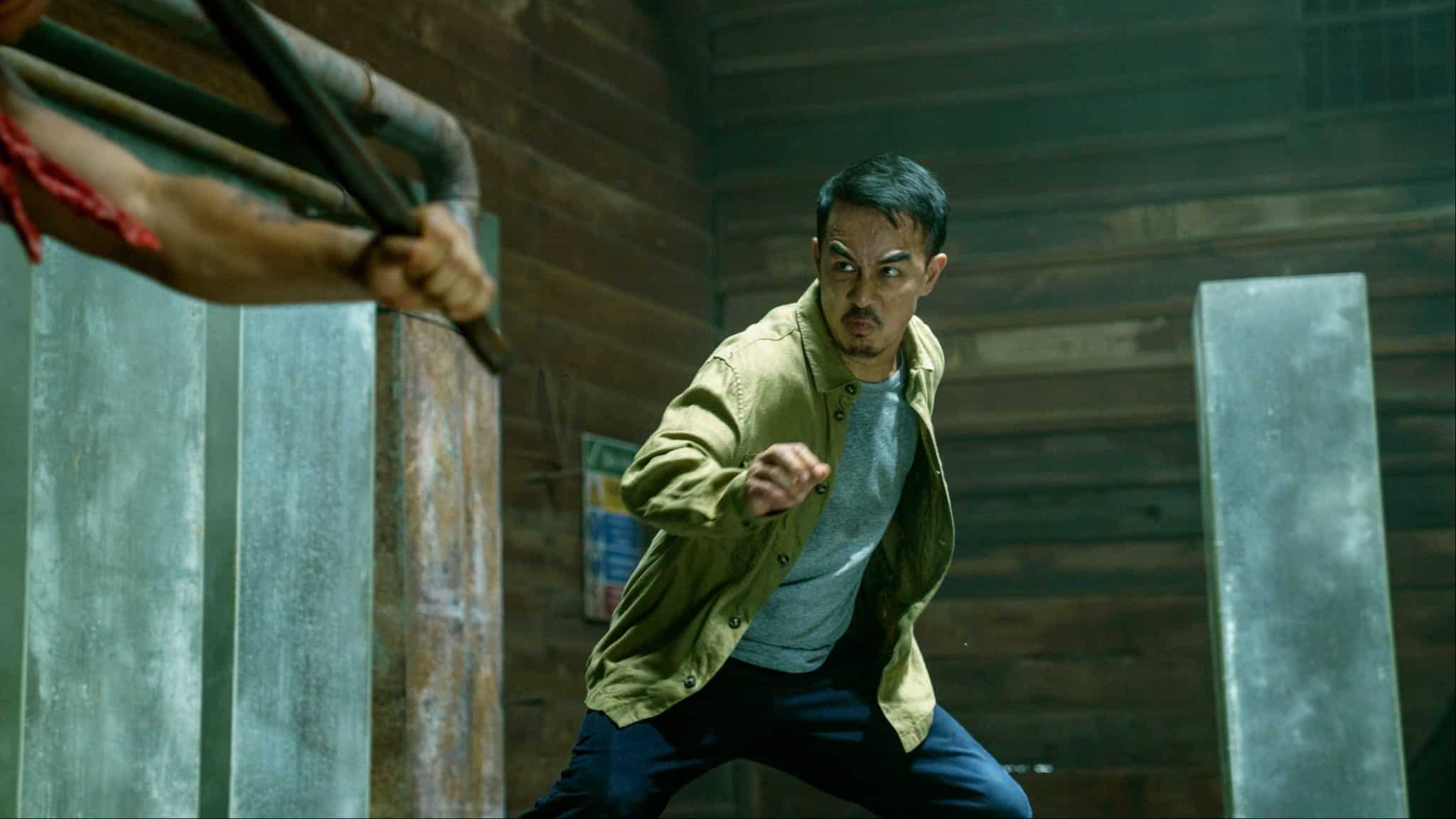 A man in a tan jacket and gray shirt clenches his fist and prepares to fight, facing another person holding a weapon in a rustic wooden room.