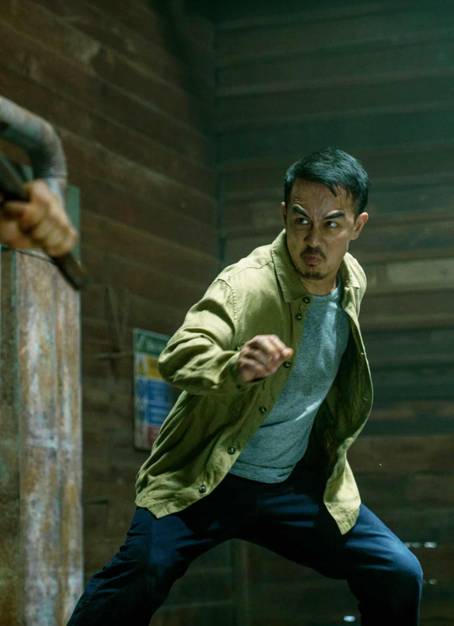 A man in a tan jacket and gray shirt clenches his fist and prepares to fight, facing another person holding a weapon in a rustic wooden room.