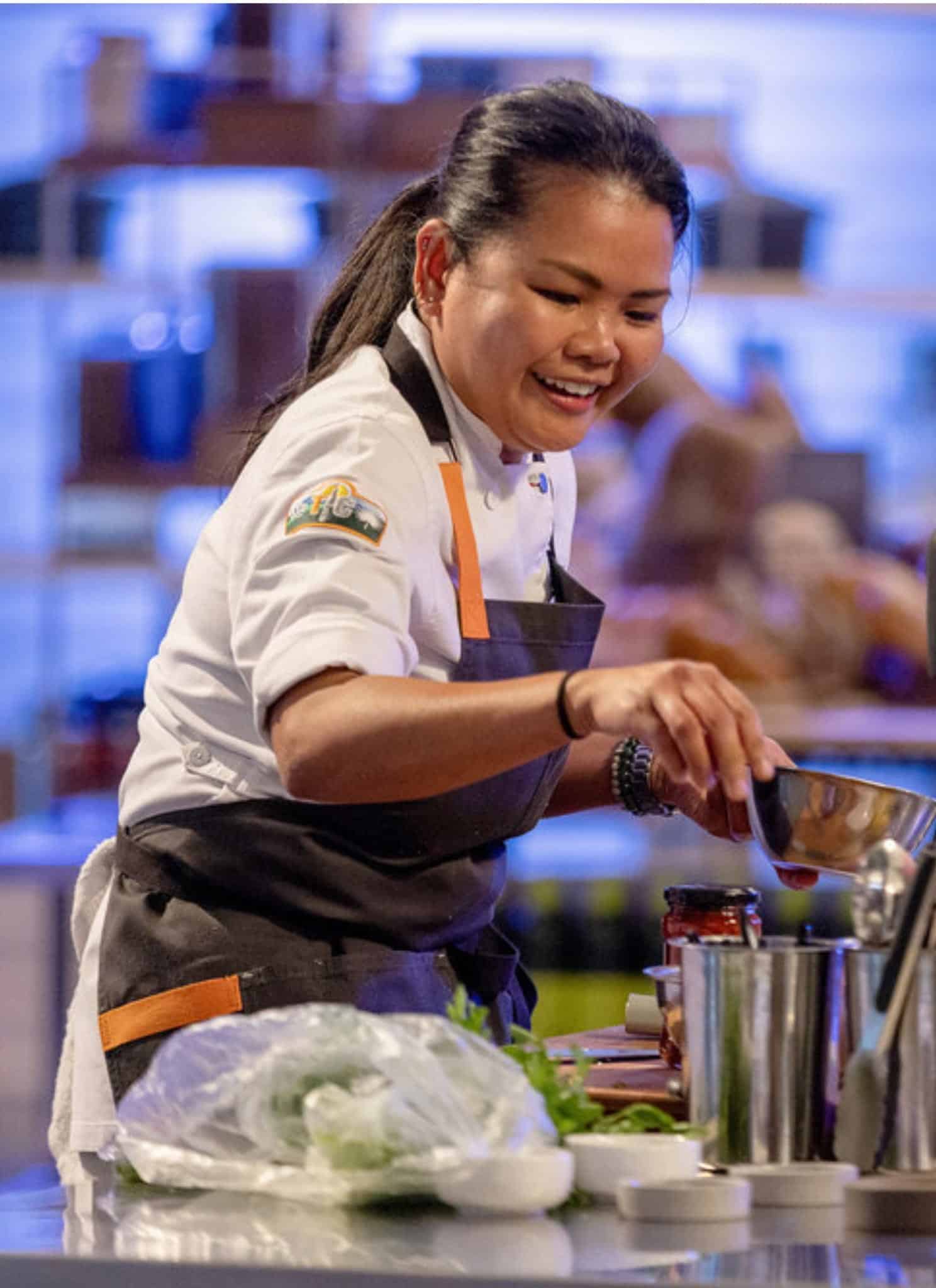 A chef in uniform smiles while preparing food at a counter filled with kitchen tools, ingredients, and a blender in a brightly lit cooking competition setting.