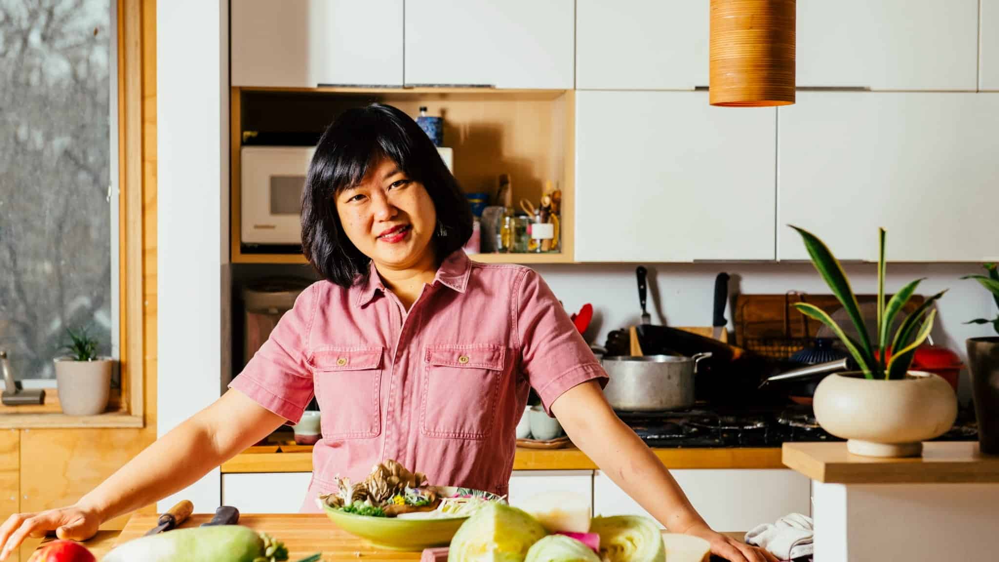 A person with shoulder-length dark hair and a pink shirt stands in a bright kitchen, smiling and leaning on a counter with fresh vegetables and a prepared dish in front of them. Cooking utensils and plants are in the background.