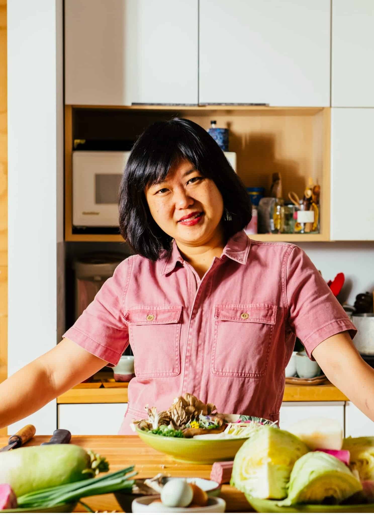 A person with shoulder-length dark hair and a pink shirt stands in a bright kitchen, smiling and leaning on a counter with fresh vegetables and a prepared dish in front of them. Cooking utensils and plants are in the background.