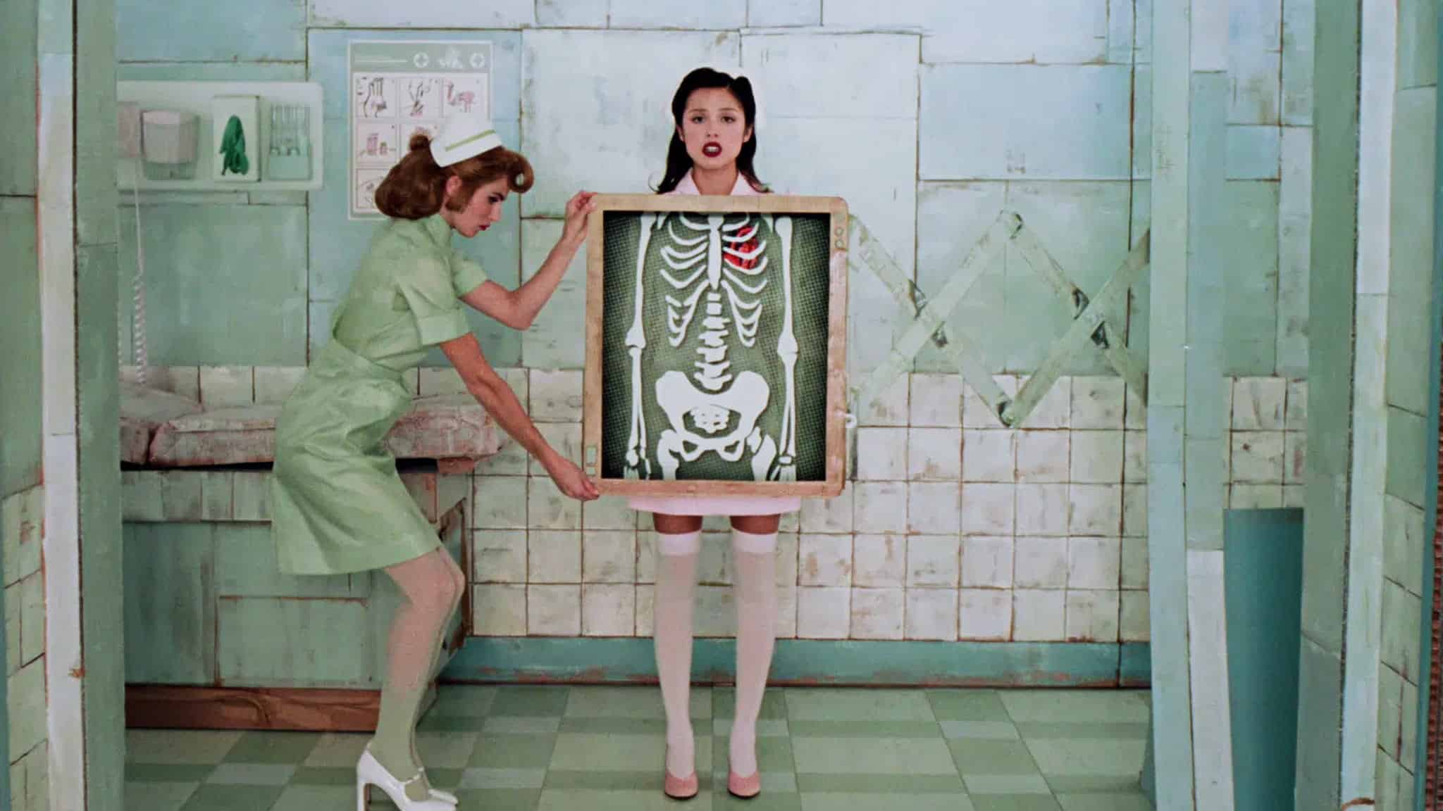 A nurse in a green uniform holds an X-ray in front of a woman in white stockings, revealing her skeleton with a red-highlighted rib. They stand in a retro, pale green-tiled medical room.