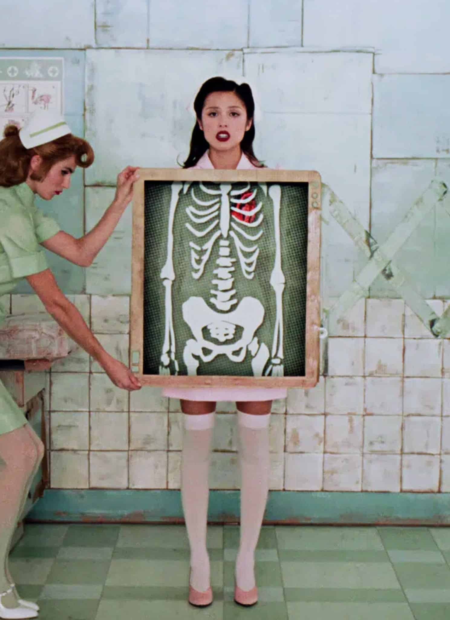 A nurse in a green uniform holds an X-ray in front of a woman in white stockings, revealing her skeleton with a red-highlighted rib. They stand in a retro, pale green-tiled medical room.