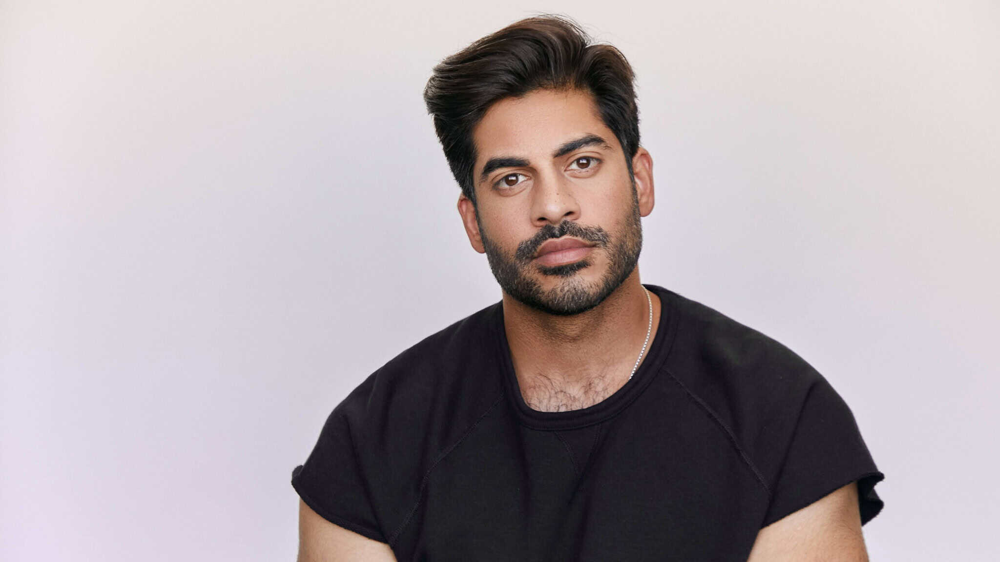 Sachin Bhatt, a man with dark hair and a beard, wearing a black t-shirt, looks calmly at the camera against a plain light background.