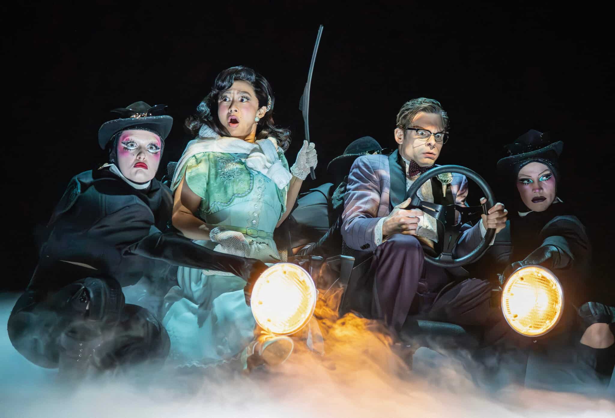 A group of actors on stage dramatize a car ride, with one person holding a steering wheel and others surrounding them, dressed in theatrical costumes with dramatic lighting and fog effects.