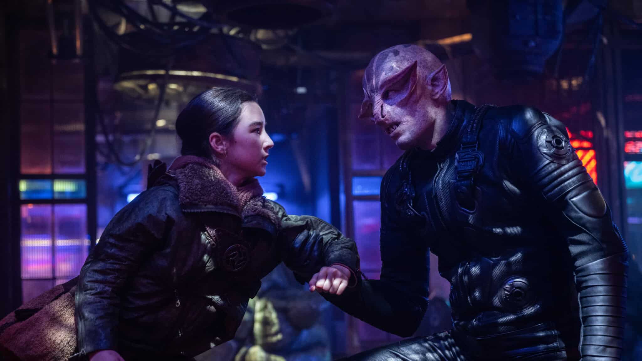 A young woman in rugged clothing grabs the arm of an alien with purple skin and pointed ears, both in a dimly lit, futuristic setting with blue and purple lights.