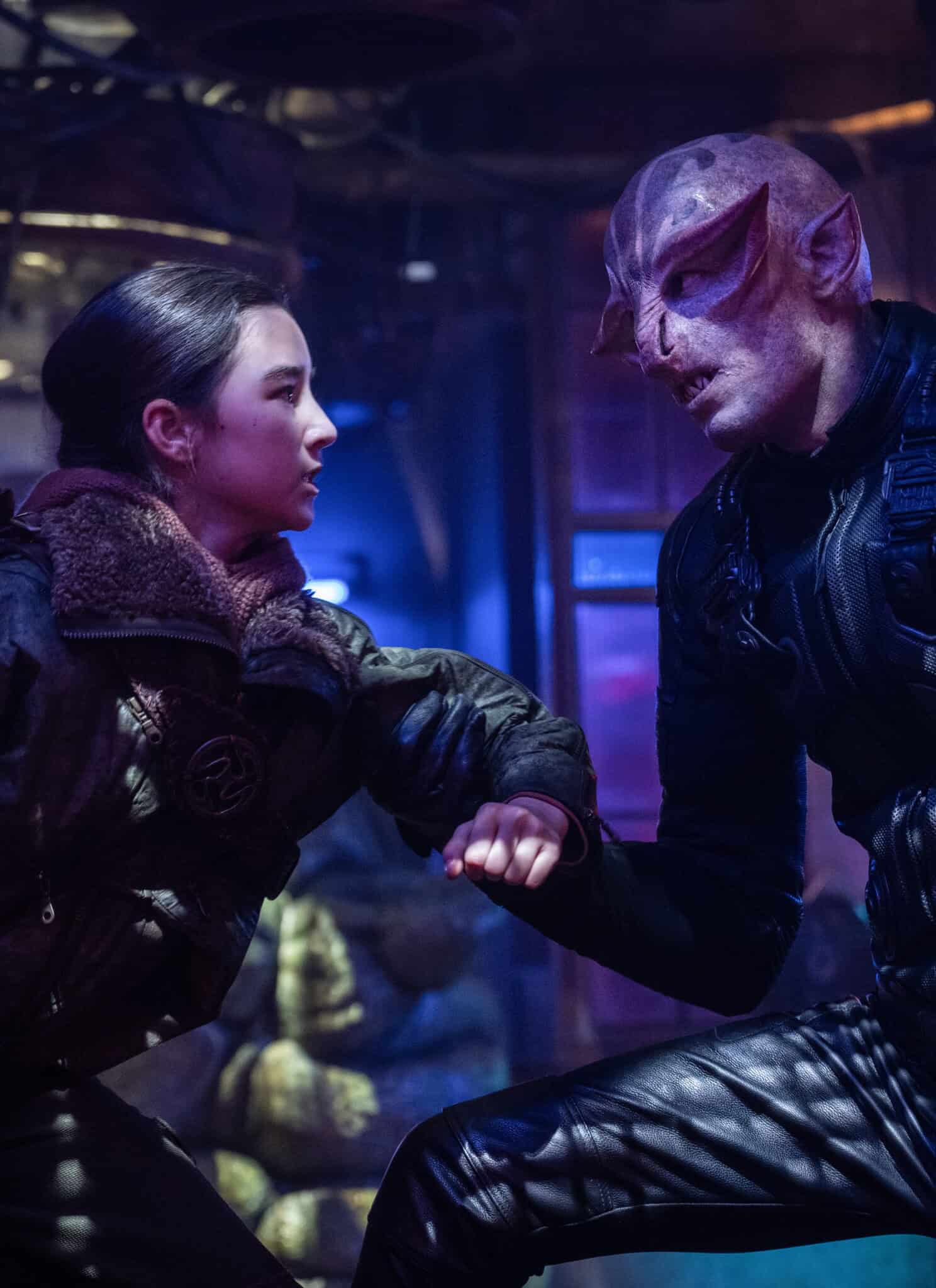 A young woman in rugged clothing grabs the arm of an alien with purple skin and pointed ears, both in a dimly lit, futuristic setting with blue and purple lights.