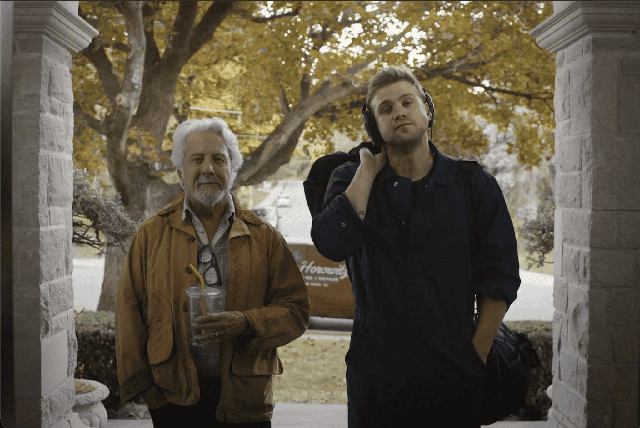 Two men stand on a porch framed by stone pillars. The older man on the left has gray hair, a beard, and wears a brown jacket, holding a drink cup. The younger man, reminiscent of Havana Rose Liu’s style, wears a dark jacket and carries a bag over his shoulder.