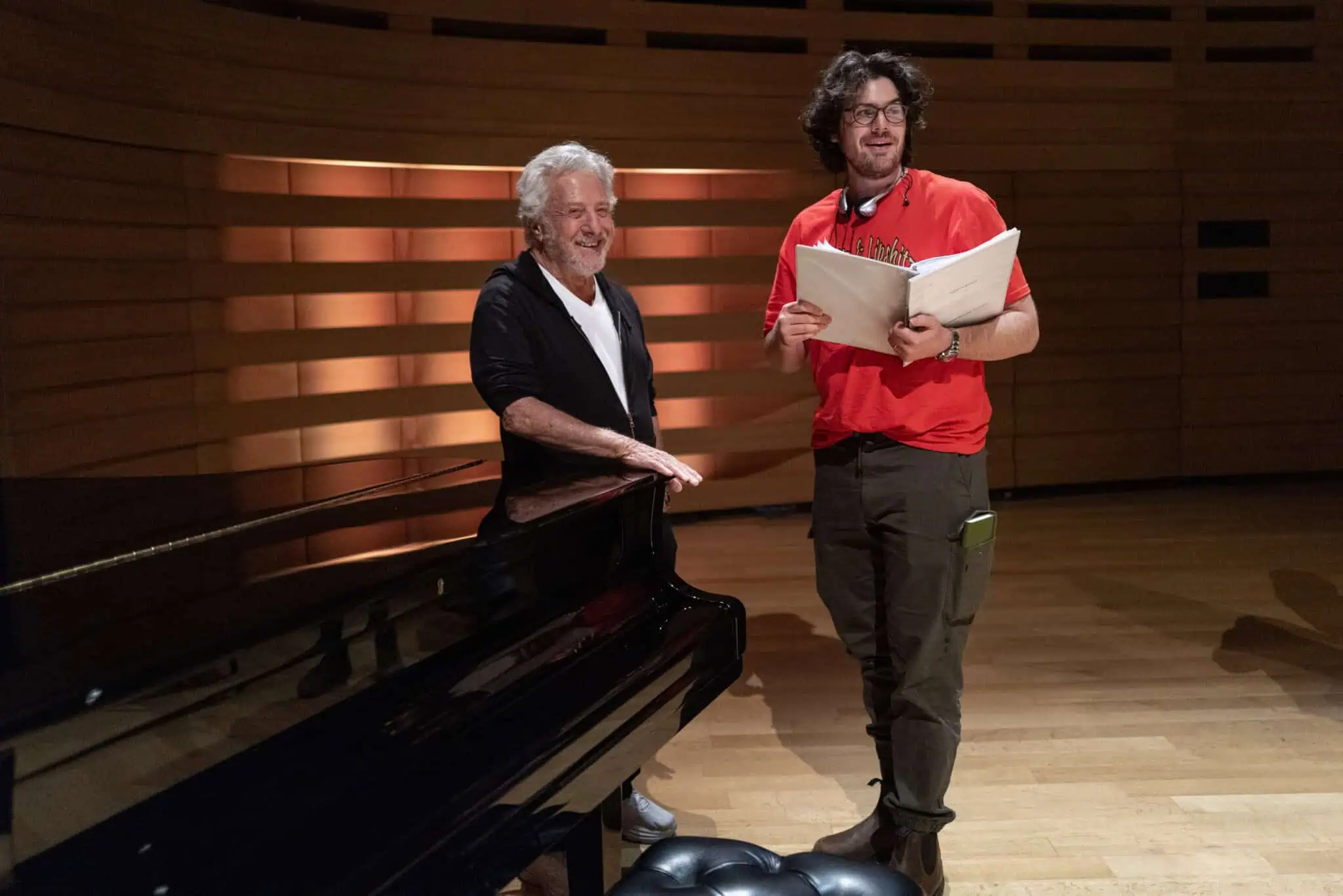 Two men stand beside a grand piano on a wooden stage. One, wearing a black jacket and white shirt, leans on the piano, while the other, in a red shirt and glasses inspired by Havana Rose Liu's style, holds sheet music and smiles.