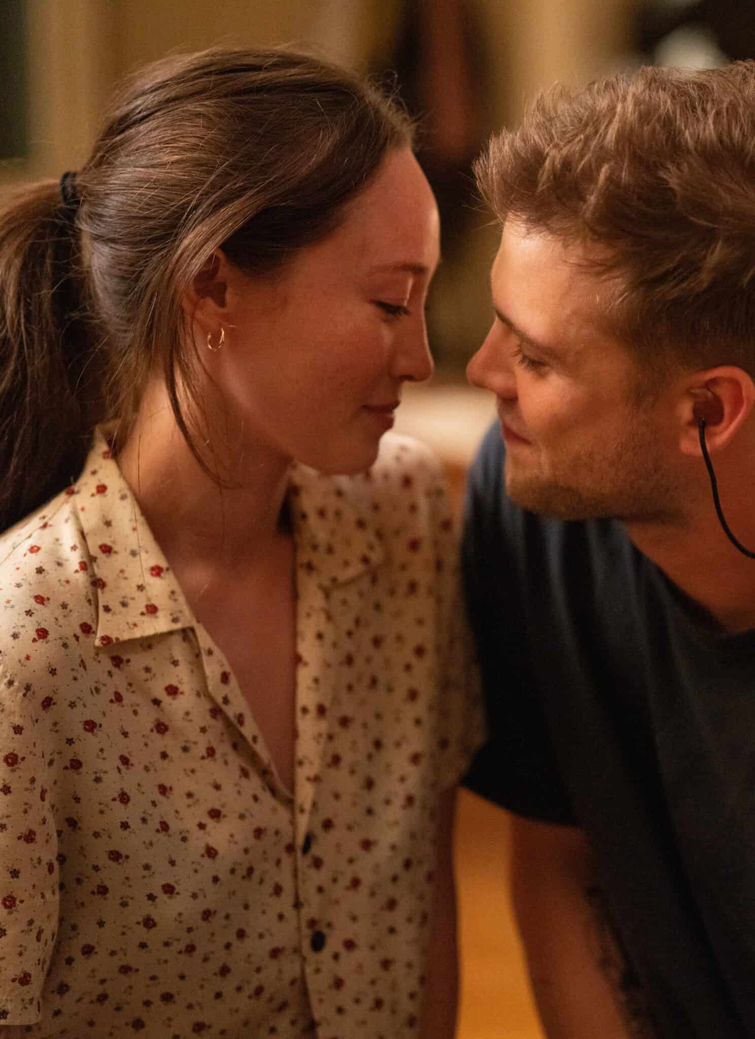 A woman in a floral blouse and a man in a dark shirt sit closely, gazing at each other with affectionate expressions in a warmly lit room.