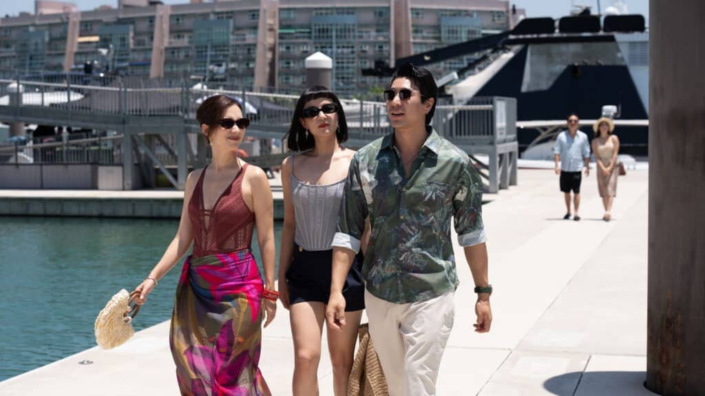 Three stylish people walk together on a sunny marina dock, smiling and wearing sunglasses, capturing the carefree spirit of The Season. A large boat and modern buildings shine in the background as two more people stroll further behind them.