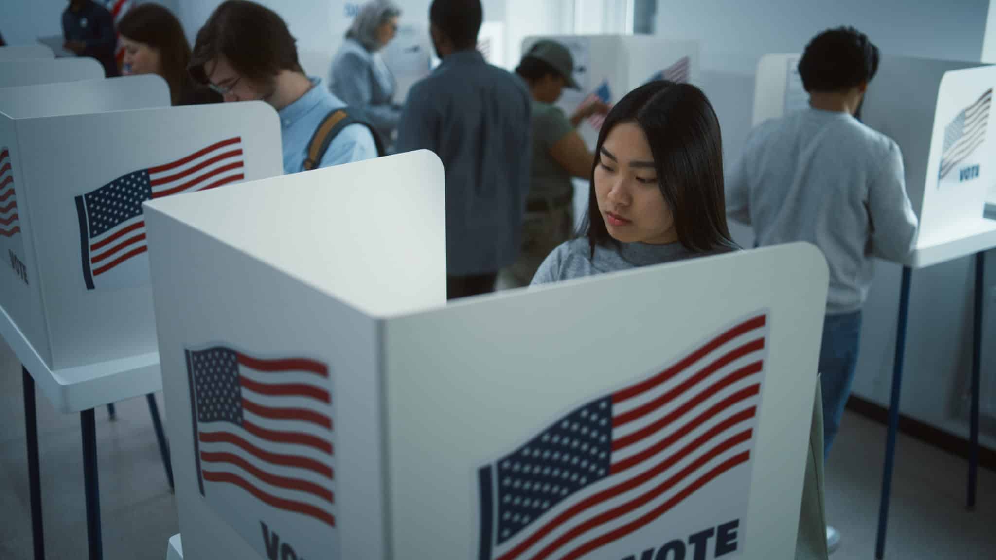 People voting in private booths at a polling station, each booth displaying an American flag and the word VOTE. The scene is diverse, with individuals focused on casting their ballots.