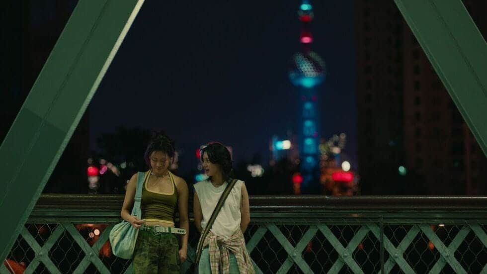 Two young women stand side by side on a bridge at night, smiling and looking down. City lights and a tall, illuminated tower are visible in the blurred background.