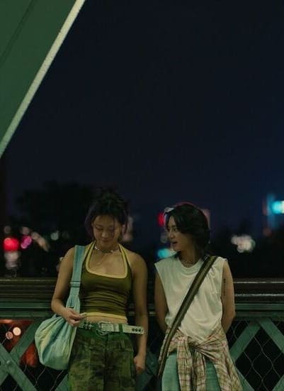Two young women stand side by side on a bridge at night, smiling and looking down. City lights and a tall, illuminated tower are visible in the blurred background.