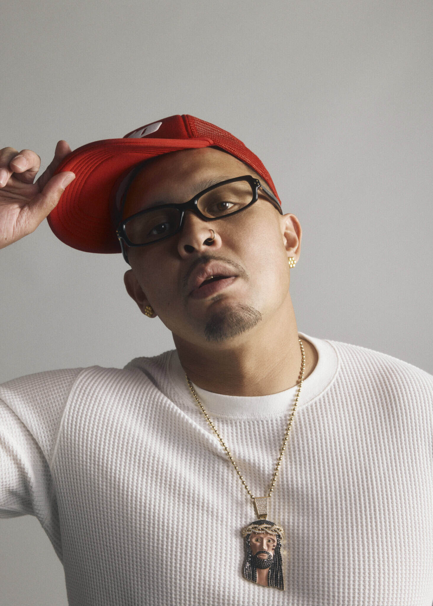 A man in a white sweater and red cap tilts his hat. He wears glasses, gold earrings, and a chain with a pendant depicting a face. The background is plain and light-colored.