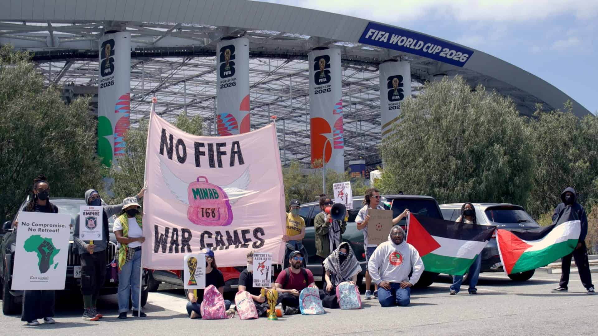 A group of people protest outside a stadium with banners reading NO FIFA WAR GAMES and NO COMPROMISE, NO RETREAT, holding signs and a Palestinian flag in front of FIFA World Cup 2026 signs.