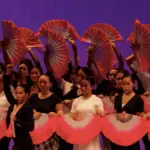 A group of dancers holds large, pink folding fans in formation on stage against a purple-lit background, wearing coordinated black and white outfits.