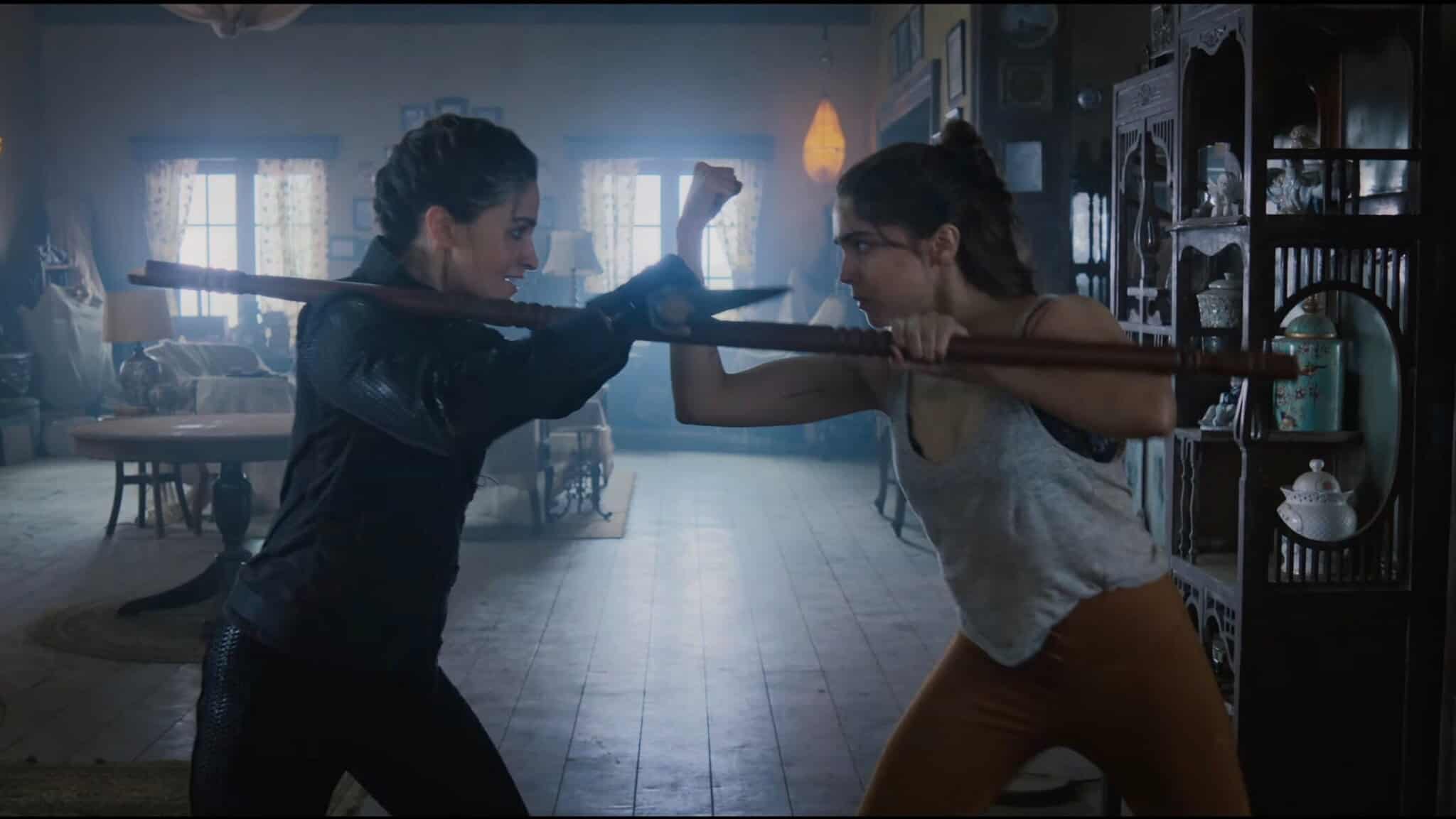 Two women face off indoors, each gripping a wooden staff and blocking each other’s moves. One wears dark clothing, the other a gray tank top and brown pants. Light streams in through the windows of the rustic room behind them.