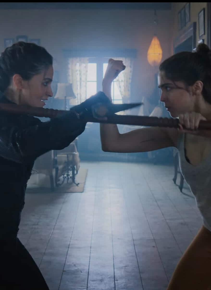Two women face off indoors, each gripping a wooden staff and blocking each other’s moves. One wears dark clothing, the other a gray tank top and brown pants. Light streams in through the windows of the rustic room behind them.