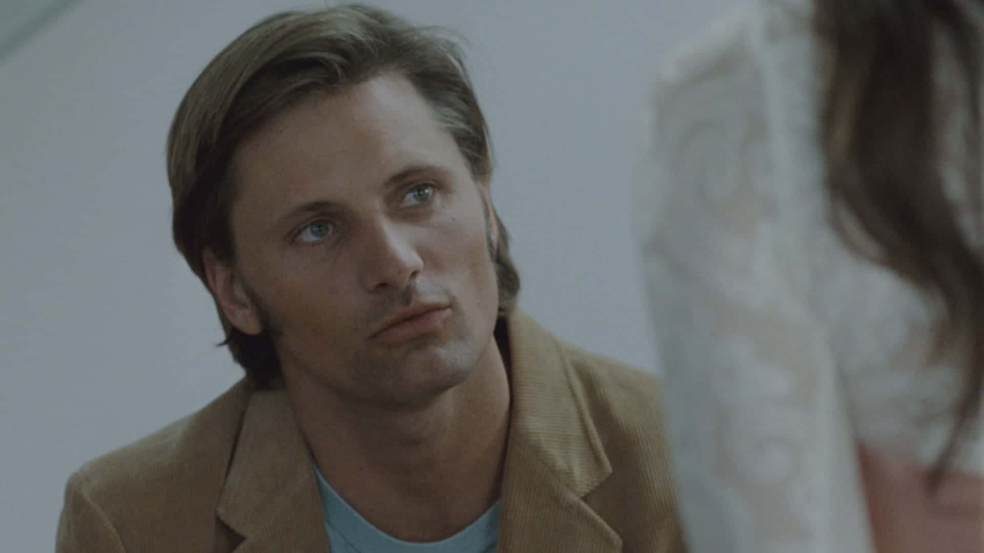 A man with light brown hair and blue eyes wears a tan jacket and blue shirt, looking intently at a person in a white, lacy garment whose face is out of focus in the foreground.