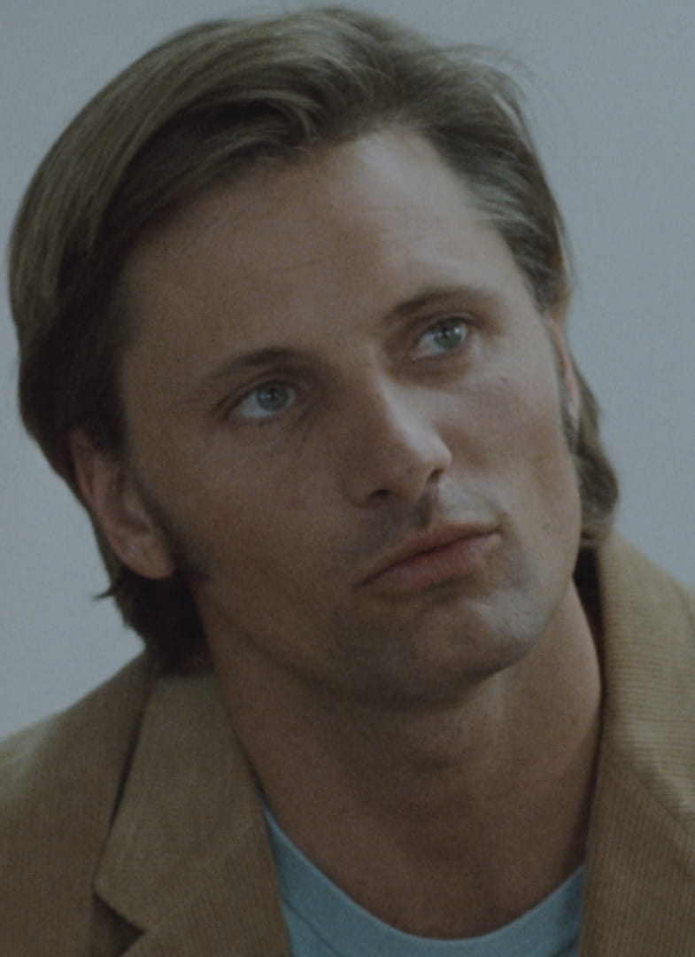 A man with light brown hair and blue eyes wears a tan jacket and blue shirt, looking intently at a person in a white, lacy garment whose face is out of focus in the foreground.