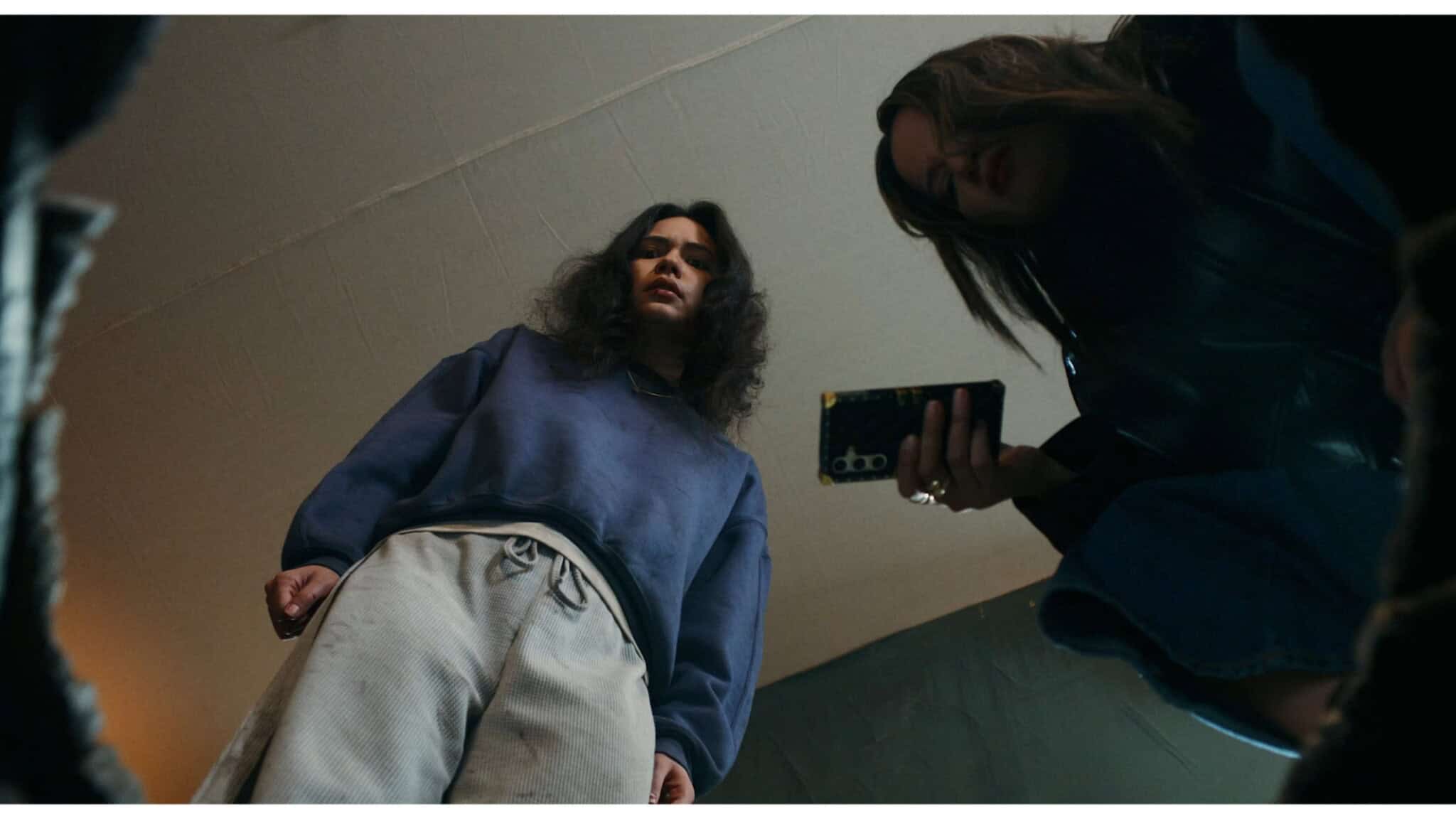 Two people look down towards the camera, one holding a phone and the other with a serious expression, both appearing to peer into a box or container. The scene is shot from a low angle.