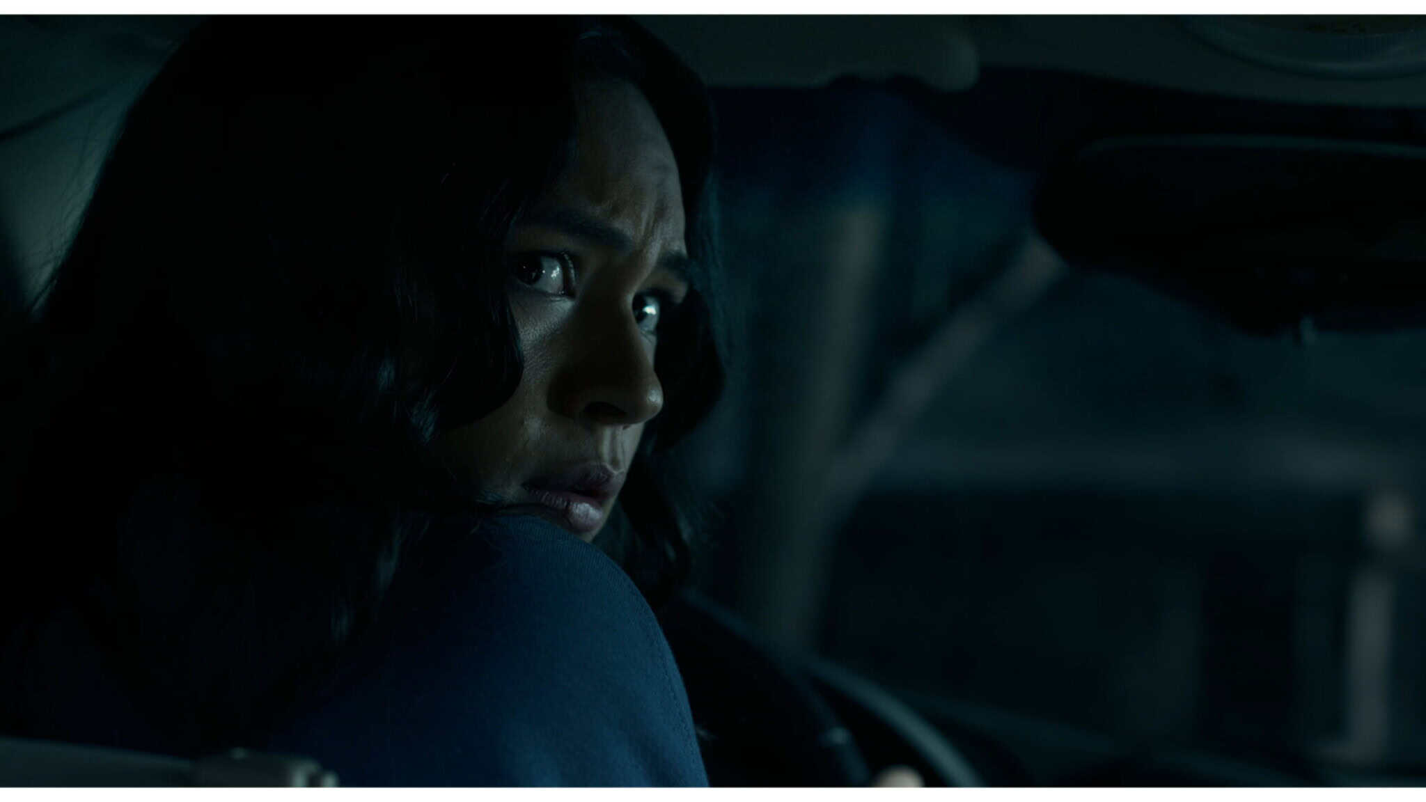 A person with long dark hair sits in a car at night, looking anxiously over their shoulder toward the back seat. The interior is dimly lit, highlighting their concerned expression.