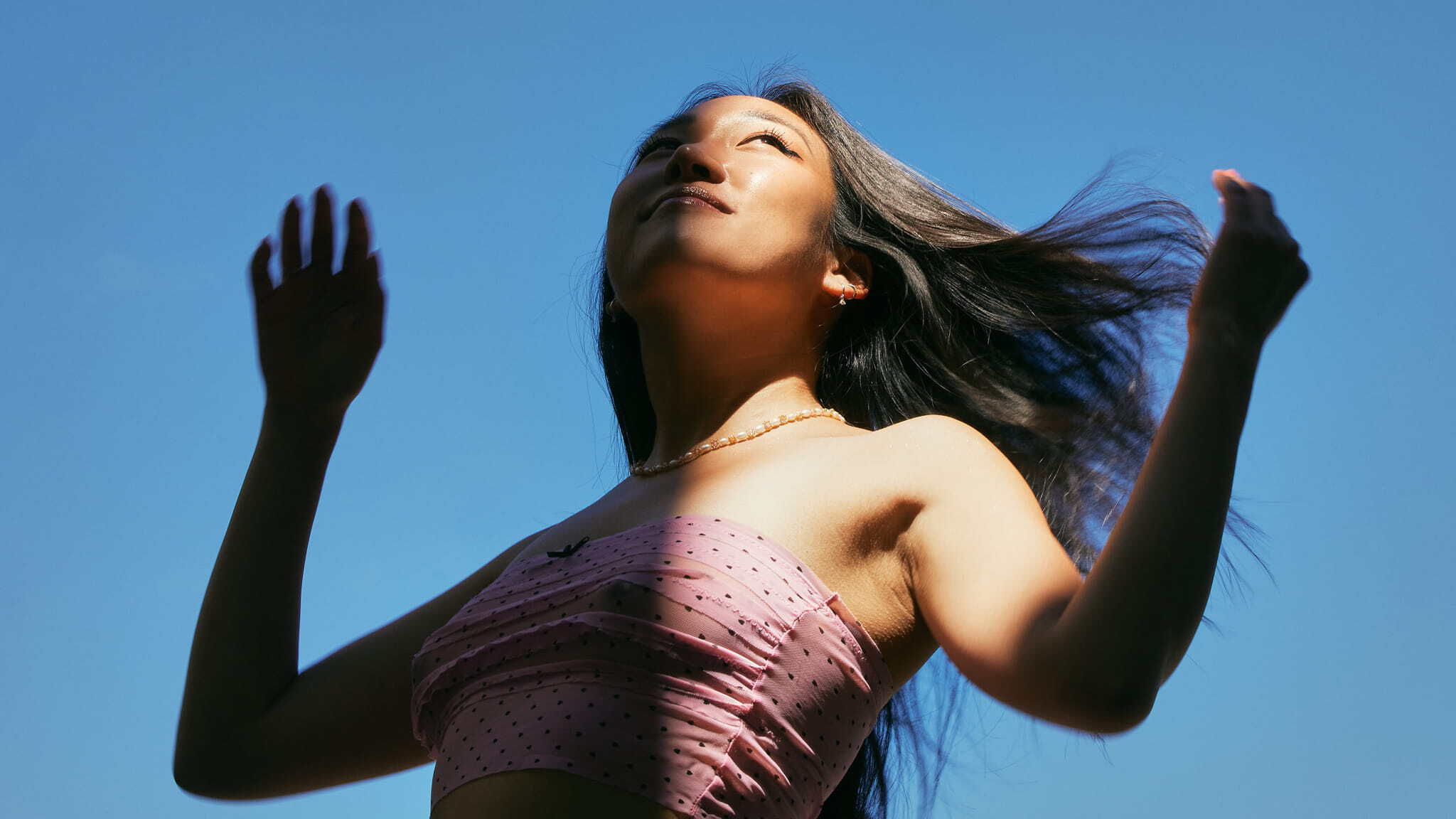 A woman in a pink strapless top stands outside against a blue sky, looking upward with her hands raised and hair blowing in the wind.