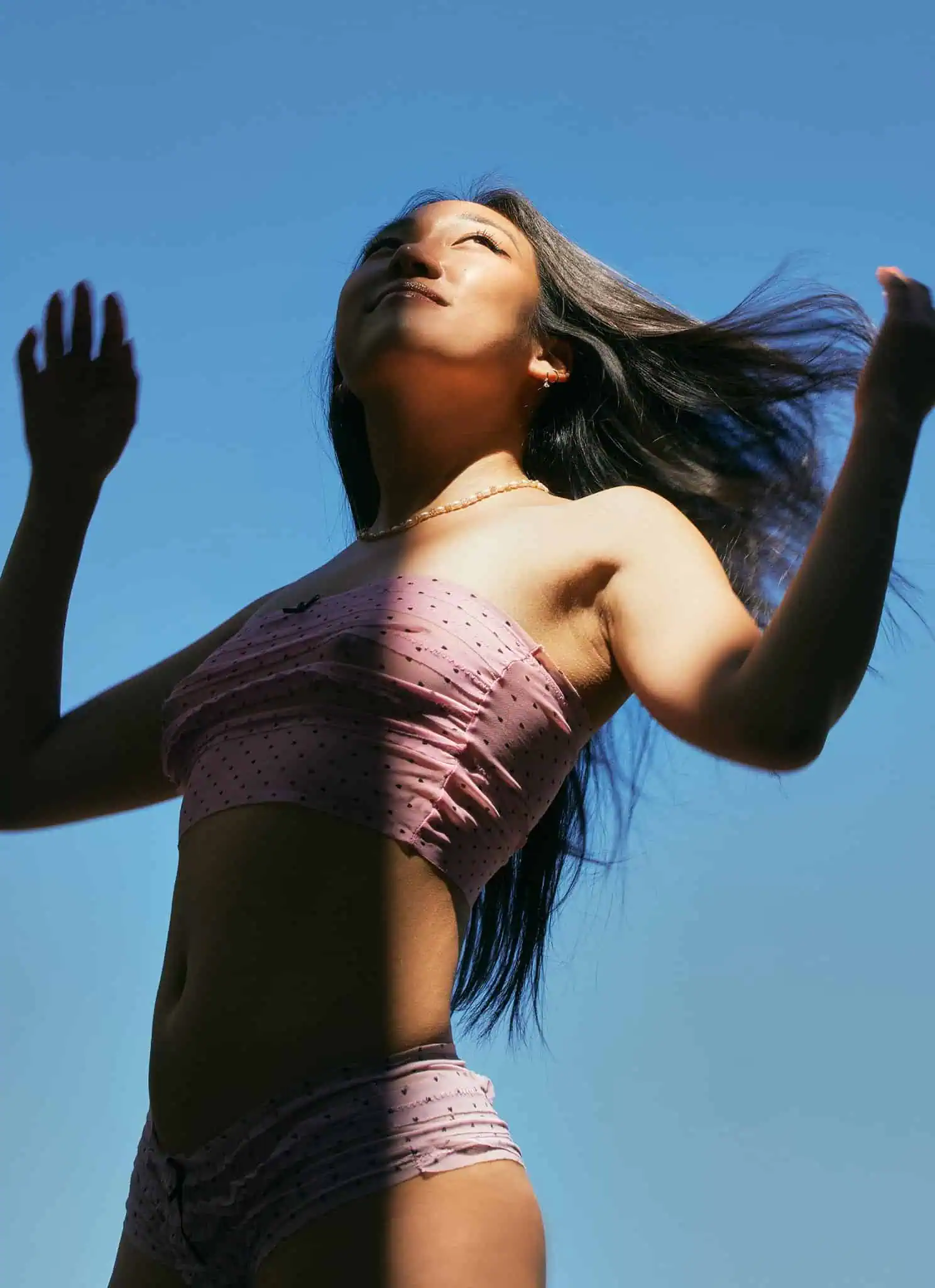 A woman in a pink strapless top stands outside against a blue sky, looking upward with her hands raised and hair blowing in the wind.
