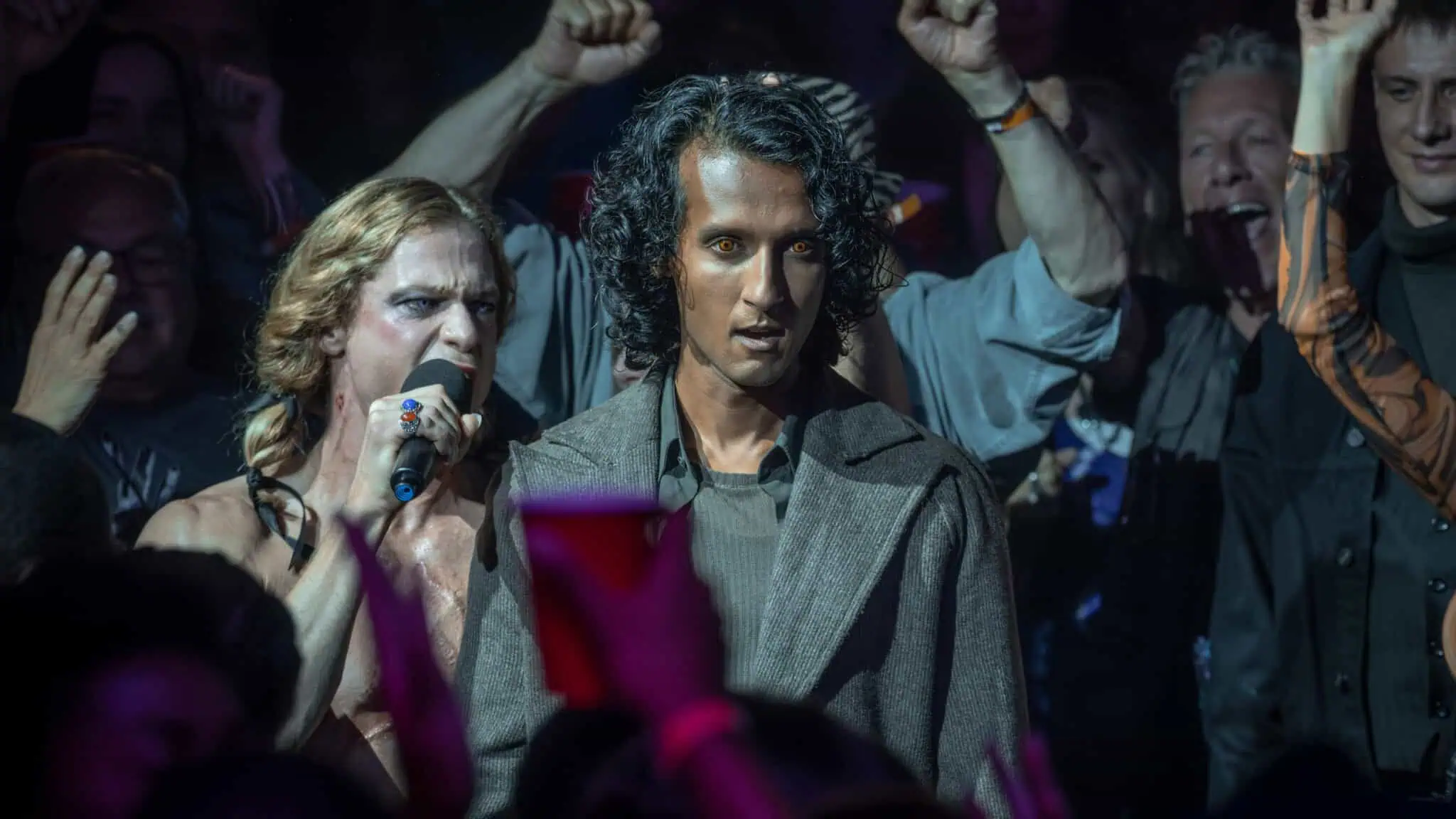 A person with curly hair and a serious expression stands in a crowd; another person sings into a microphone beside them, while people behind cheer, some raising their fists and holding red cups.