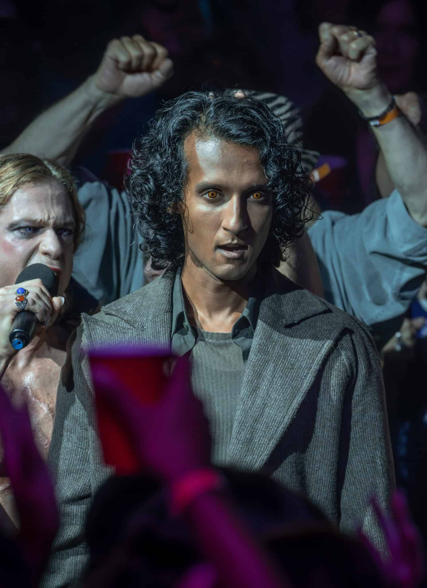A person with curly hair and a serious expression stands in a crowd; another person sings into a microphone beside them, while people behind cheer, some raising their fists and holding red cups.