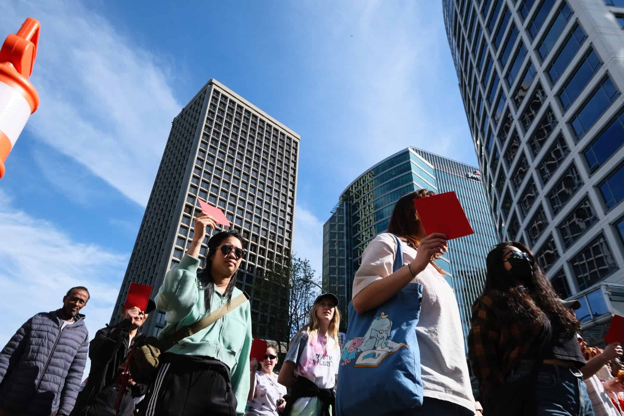 A group of people stand outdoors in a city, holding up red cards. Tall modern buildings surround them, and the sky is blue with wispy clouds.