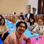 A group of people in traditional attire sit at decorated tables with small flags, smiling at the camera. A man stands behind them. The room is adorned with international flags and cultural decorations.