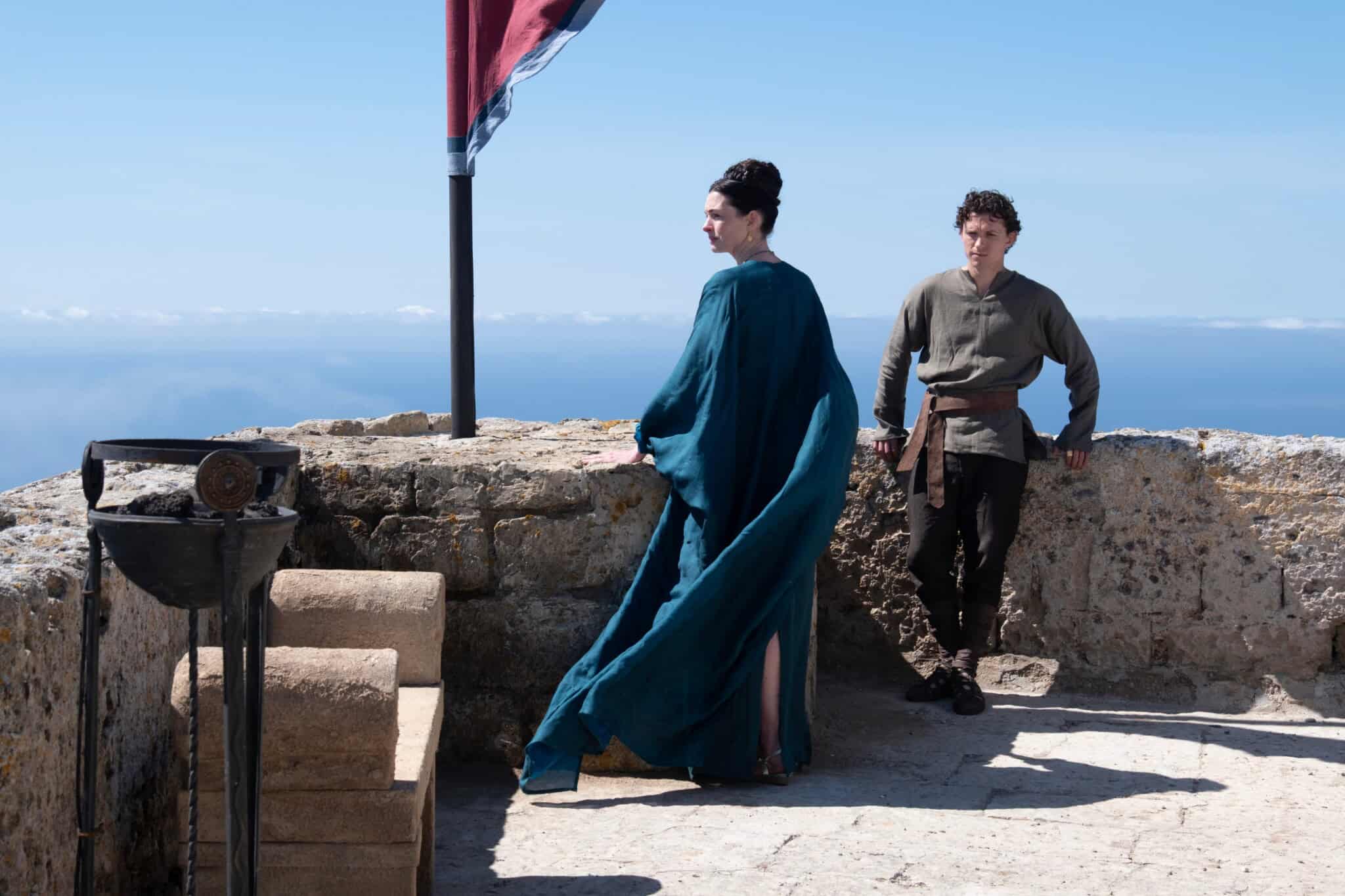 A woman in a flowing blue robe stands by a stone wall overlooking the sea, evoking scenes from The Odyssey, while a man in medieval attire stands nearby. A flagpole and a metal brazier are also visible under a clear blue sky.