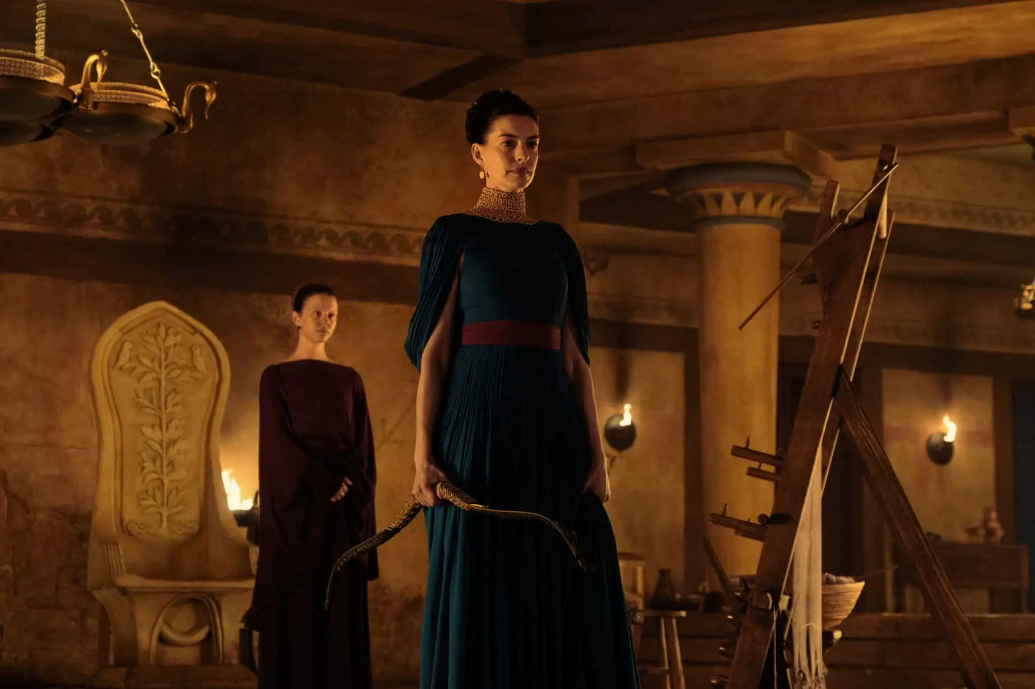 A woman in a blue dress stands holding a curved object, possibly a bow, in an ornate room with columns. Another woman in a dark dress stands behind her. The setting is lit by warm, ambient lighting.