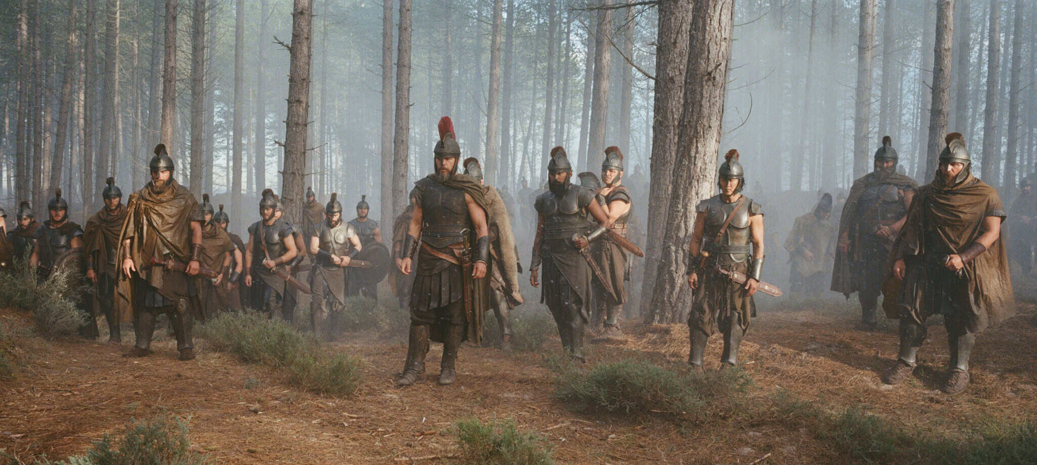 A group of ancient soldiers in armor and helmets stands in a misty forest, some holding swords and shields, with tall trees and scattered bushes around them.
