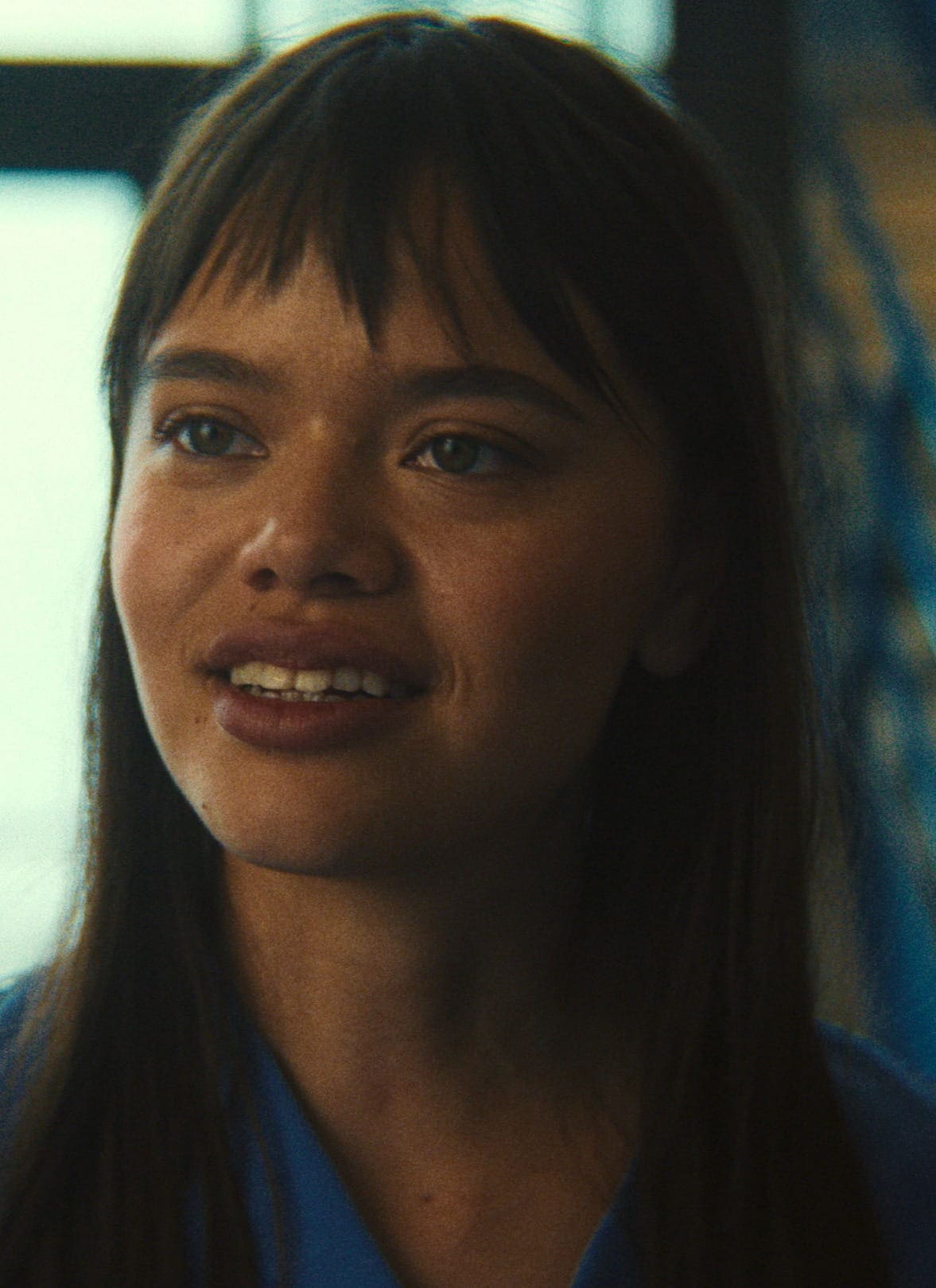 A young woman with straight brown hair and bangs smiles while talking to someone whose back is facing the camera. The background is softly lit, with some blue and yellow tones.