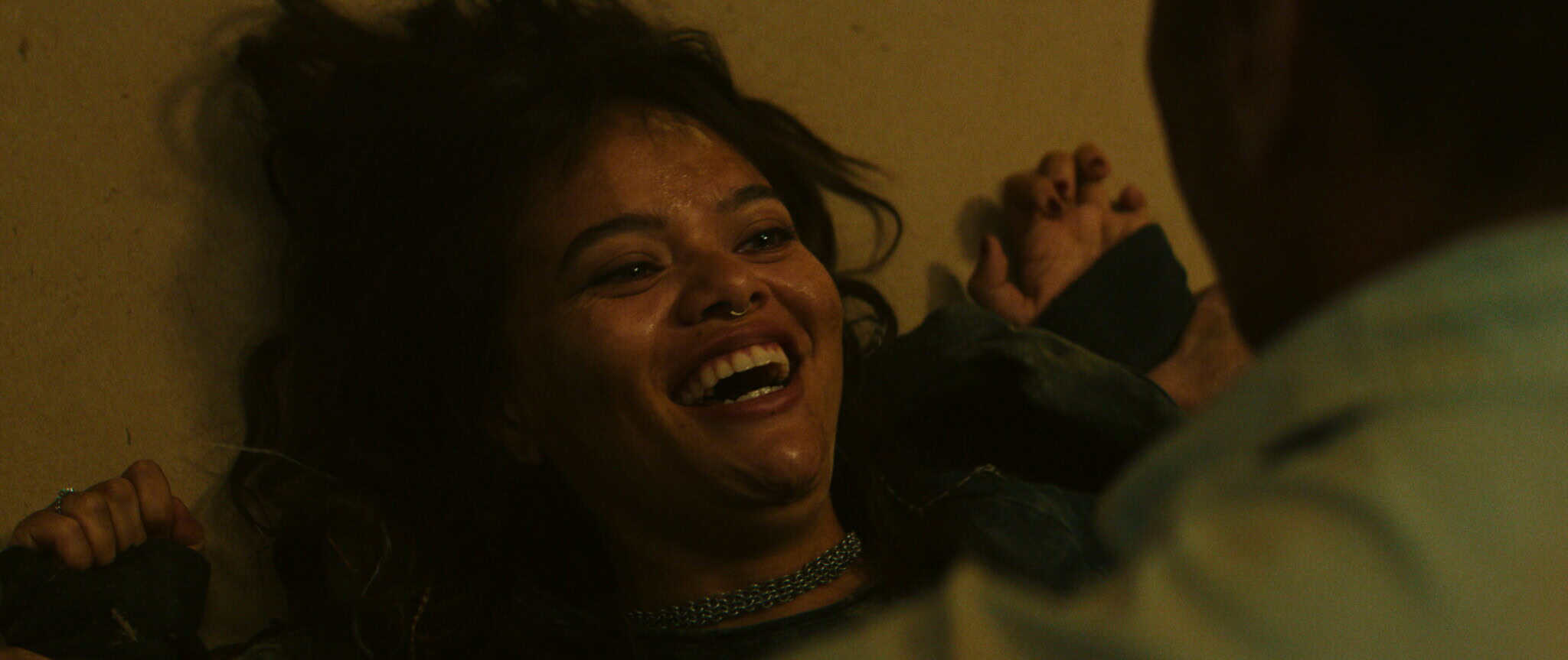 A woman with long dark hair lies on the floor, smiling and laughing, while a person in a white shirt is partially visible facing her in the foreground.