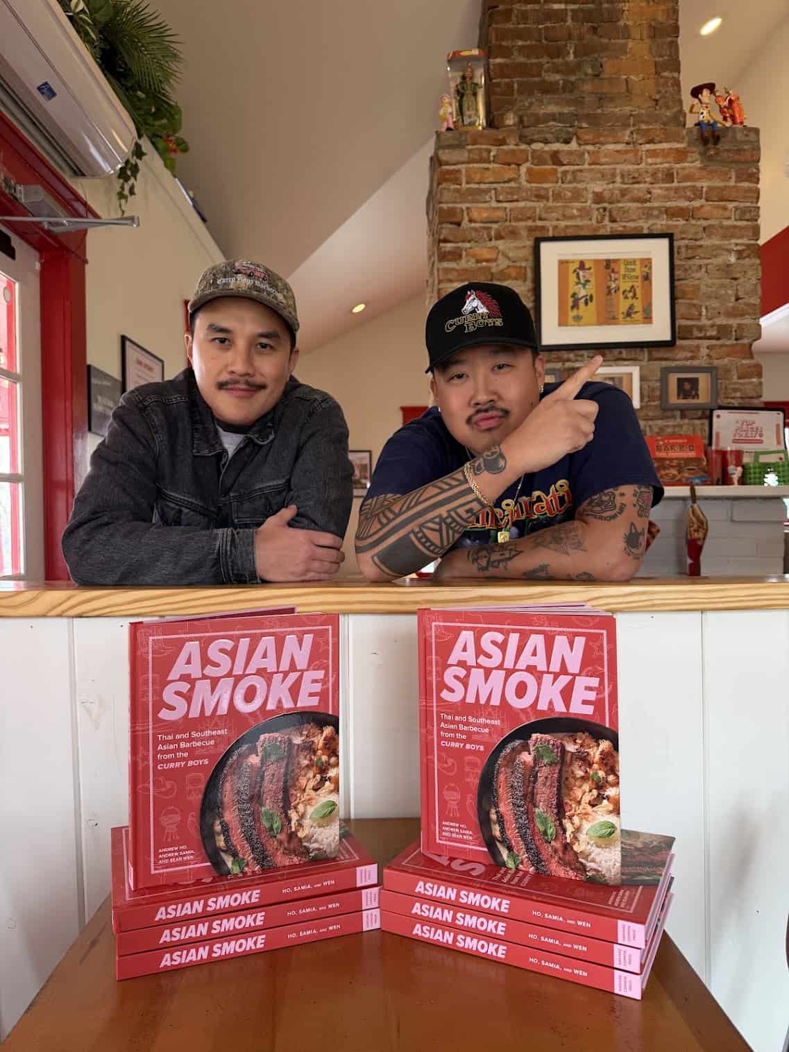 Two men pose behind a counter with several copies of a cookbook titled Asian Smoke. The background features a brick wall, framed art, and shelves with various items.