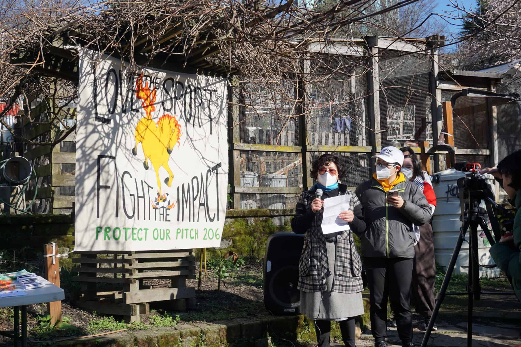 Two people stand outdoors near a large protest sign that reads, “FIGHT THE IMPACT.” One speaks into a microphone while the other holds papers. A person films the scene. Leafless branches and a wooden structure are visible behind them.