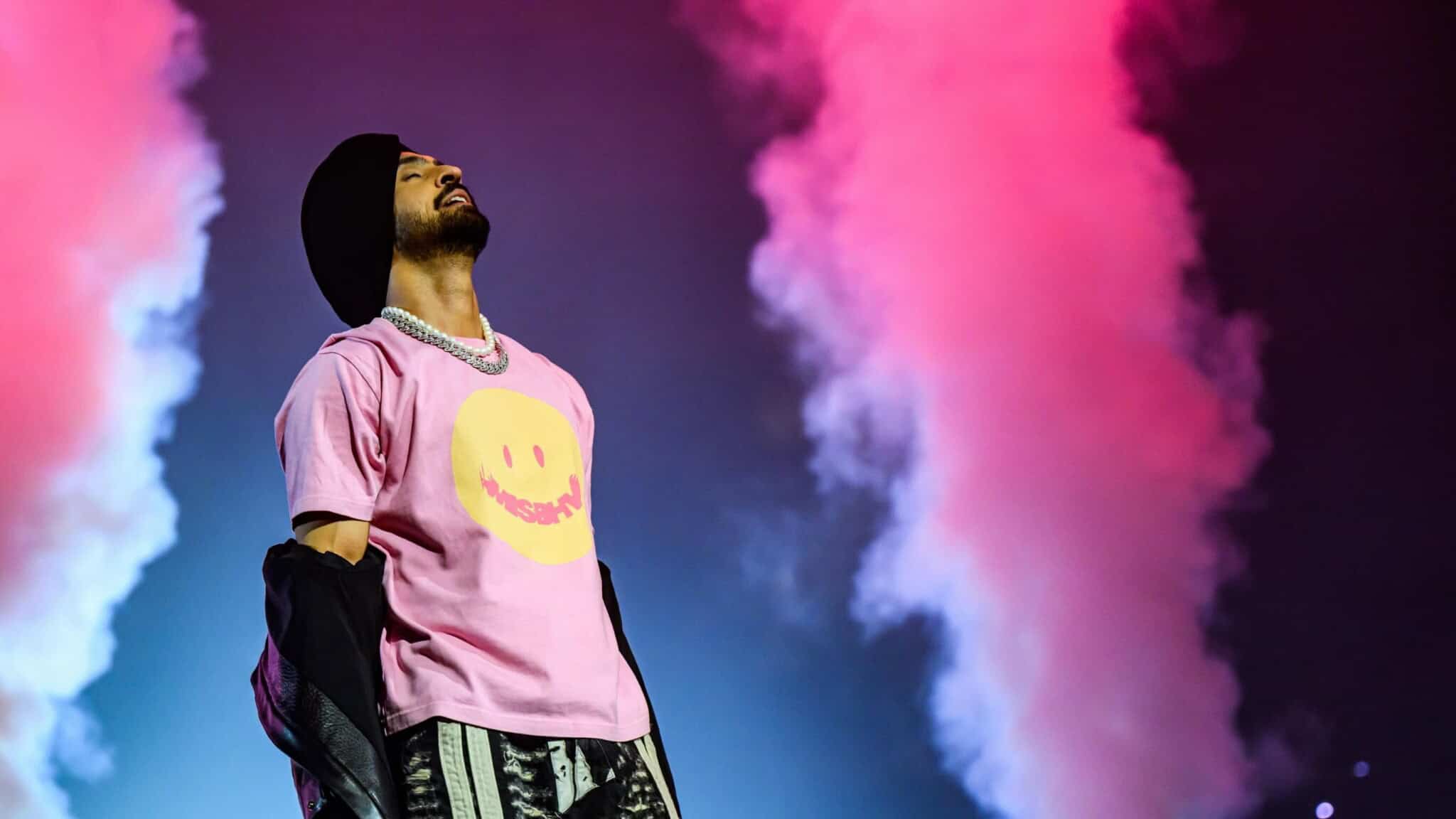 A performer in a pink smiley-face shirt and black turban stands on stage with head tilted back, surrounded by pink smoke and dramatic lighting.