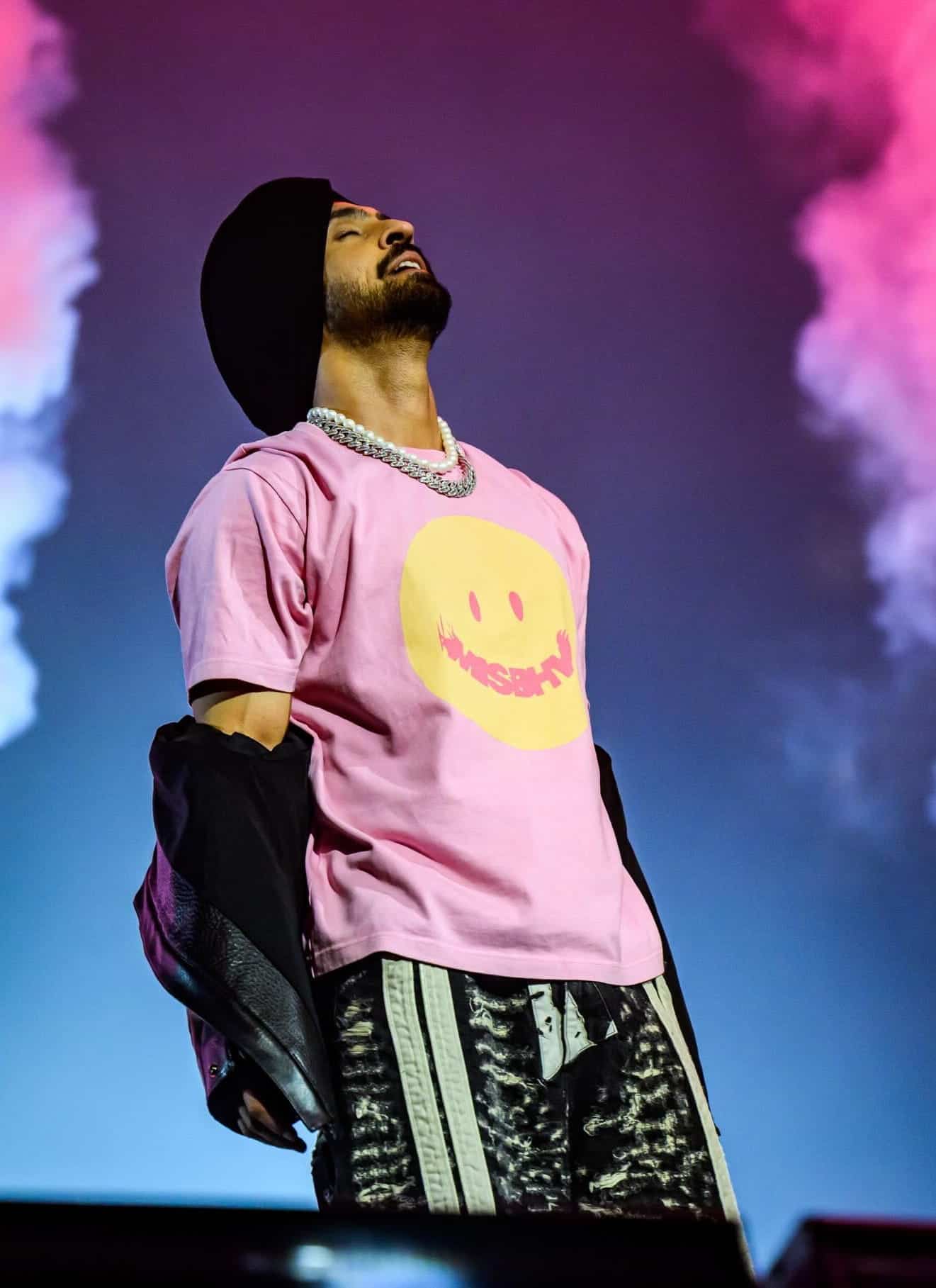 A performer in a pink smiley-face shirt and black turban stands on stage with head tilted back, surrounded by pink smoke and dramatic lighting.