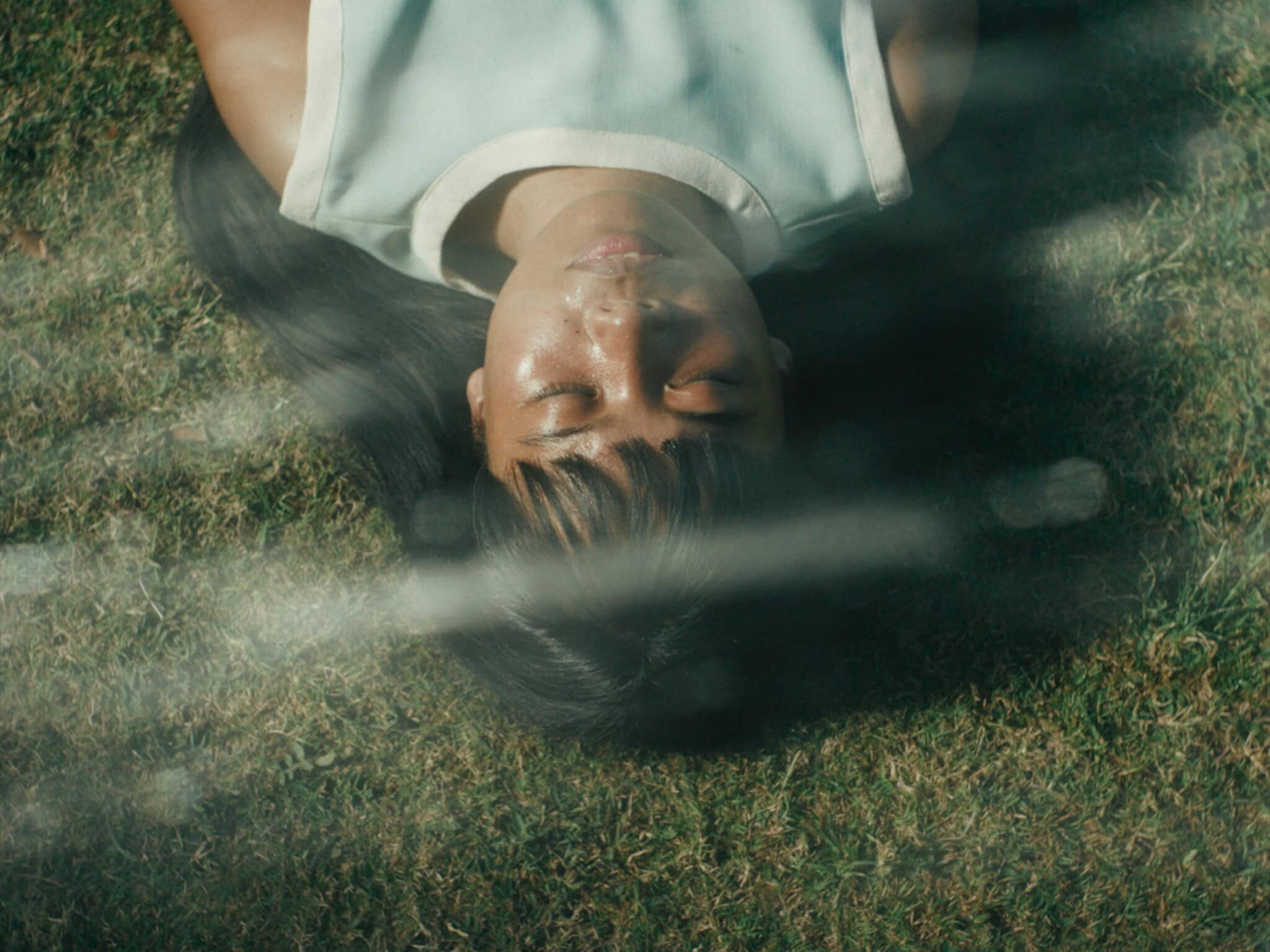 A person with long dark hair lies on grass with eyes closed, sunlight and shadows crossing their face, creating a serene and peaceful atmosphere.