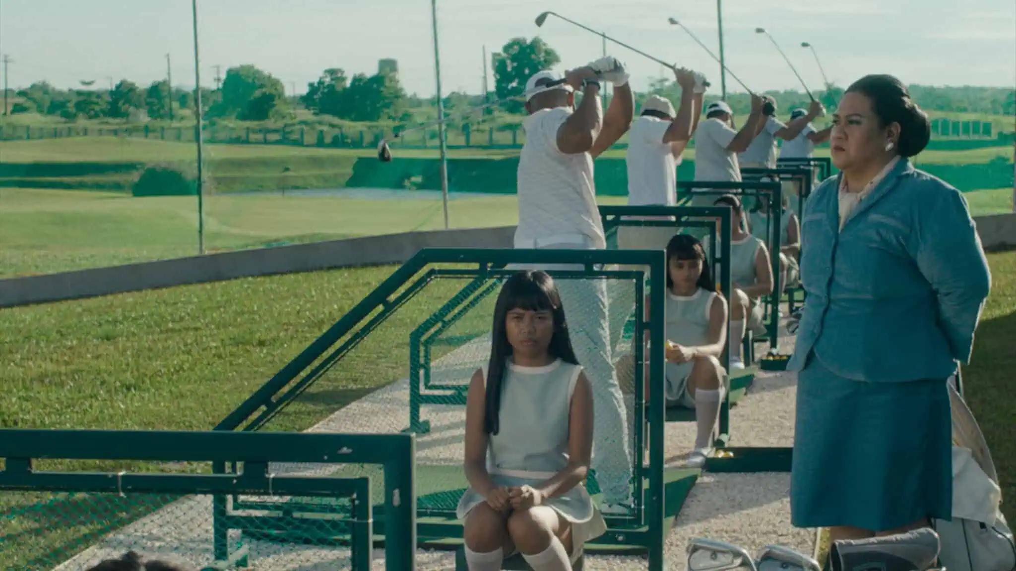 Four men in white golf outfits practice swinging at a driving range. Two girls in white dresses sit in front, and a woman in blue stands nearby. Green grass and golf course are visible in the background.