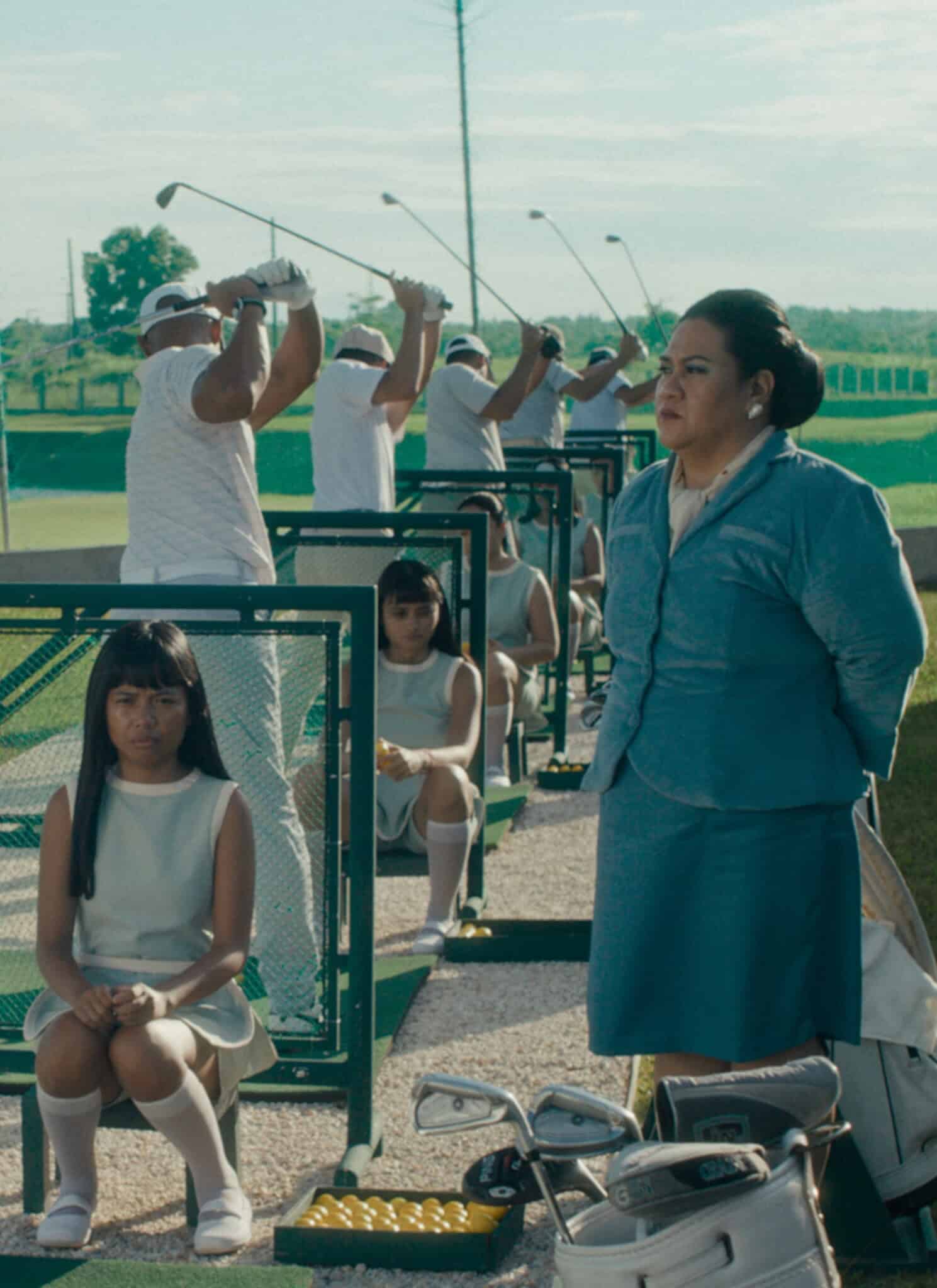 Four men in white golf outfits practice swinging at a driving range. Two girls in white dresses sit in front, and a woman in blue stands nearby. Green grass and golf course are visible in the background.
