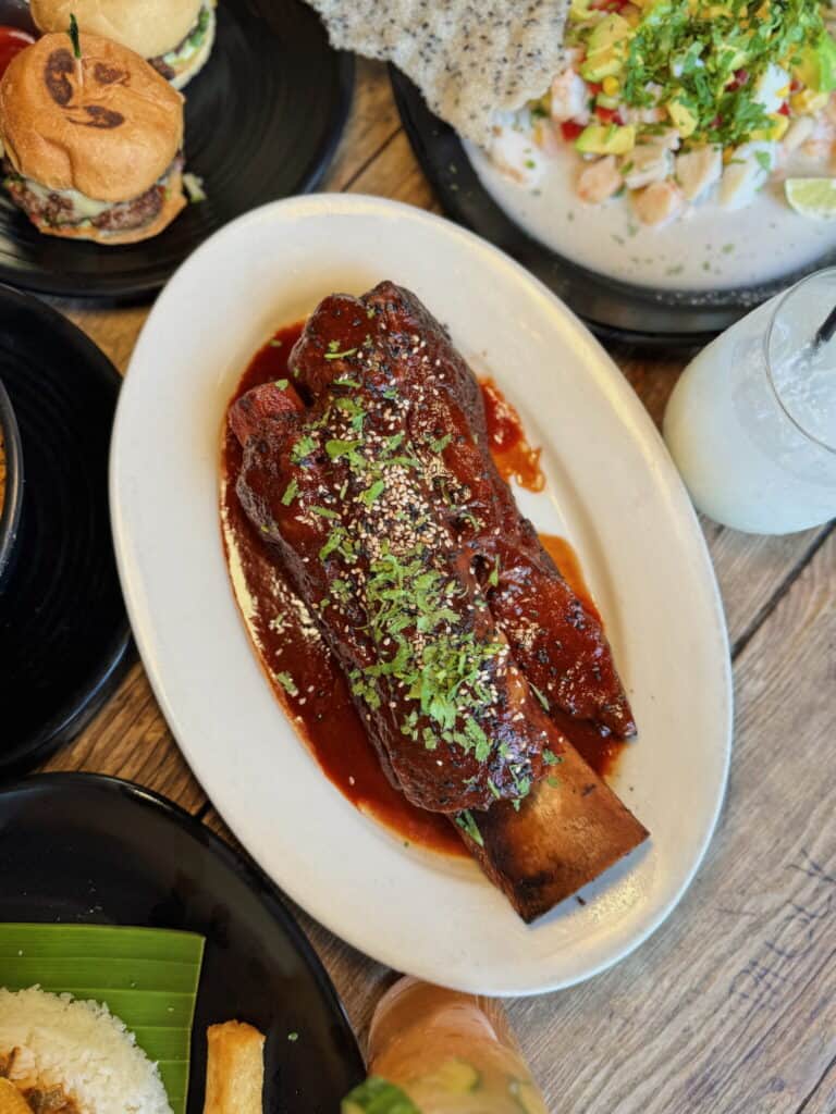 A large, saucy beef rib garnished with herbs on a white oval plate, surrounded by burgers, salad, and drinks on a wooden table.