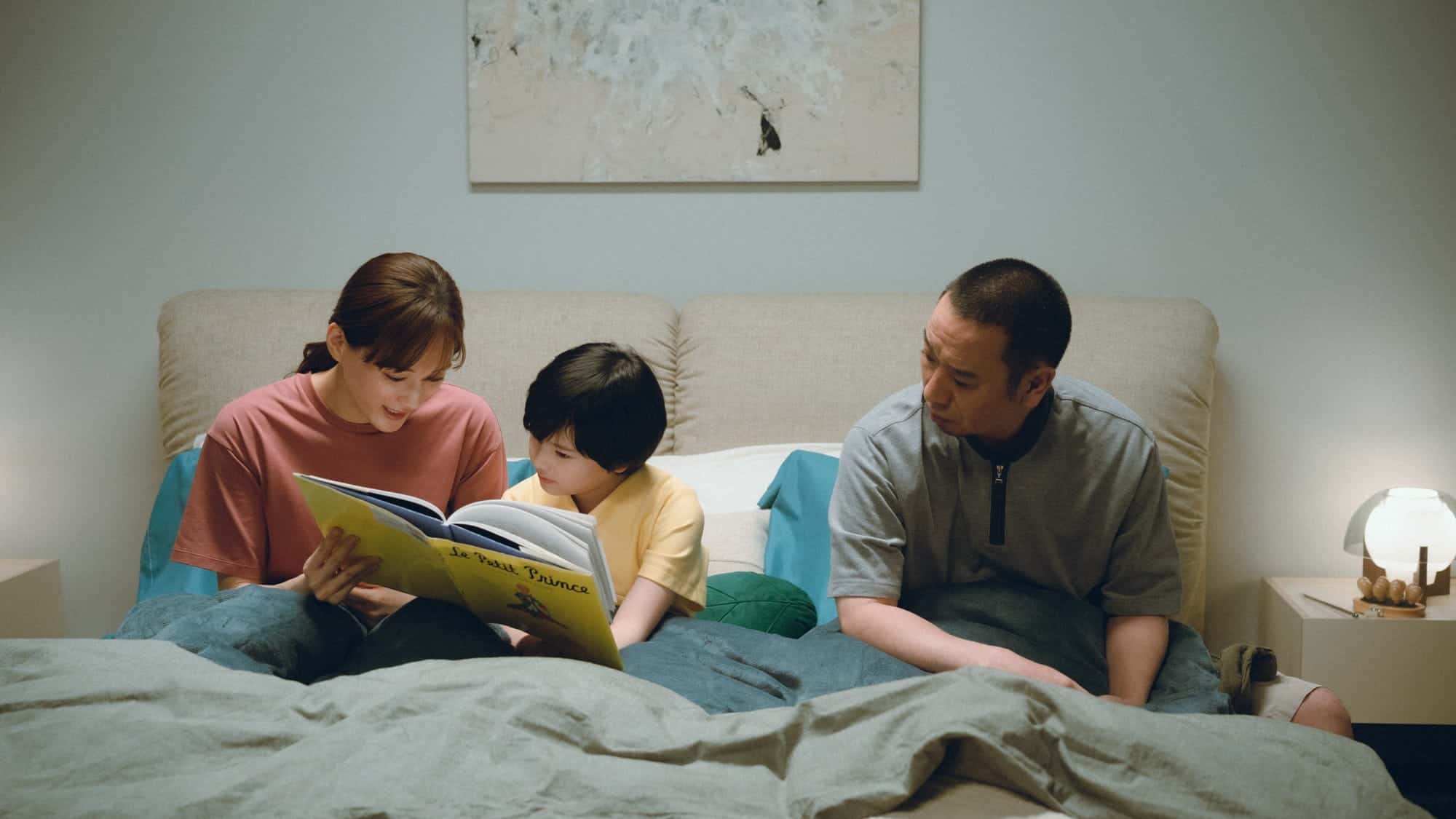 A woman reads a book to a child while a man sits beside them on a bed. They are under blankets in a softly lit bedroom with a painting on the wall and a lamp on a nightstand.