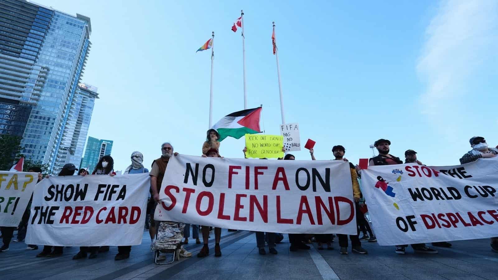 A group of protesters holds banners reading “NO FIFA ON STOLEN LAND” and “SHOW FIFA THE RED CARD,” with flags and tall buildings in the background.