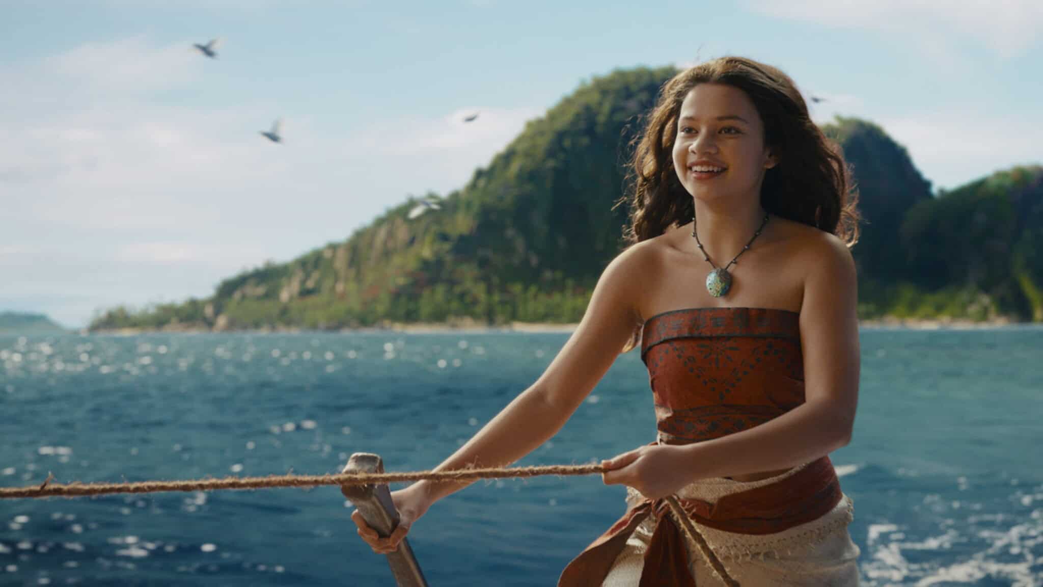 A young woman smiles while standing on a boat, holding a rope. She wears a red patterned top and a necklace. Behind her, the ocean sparkles and a green, mountainous island rises under a blue sky.