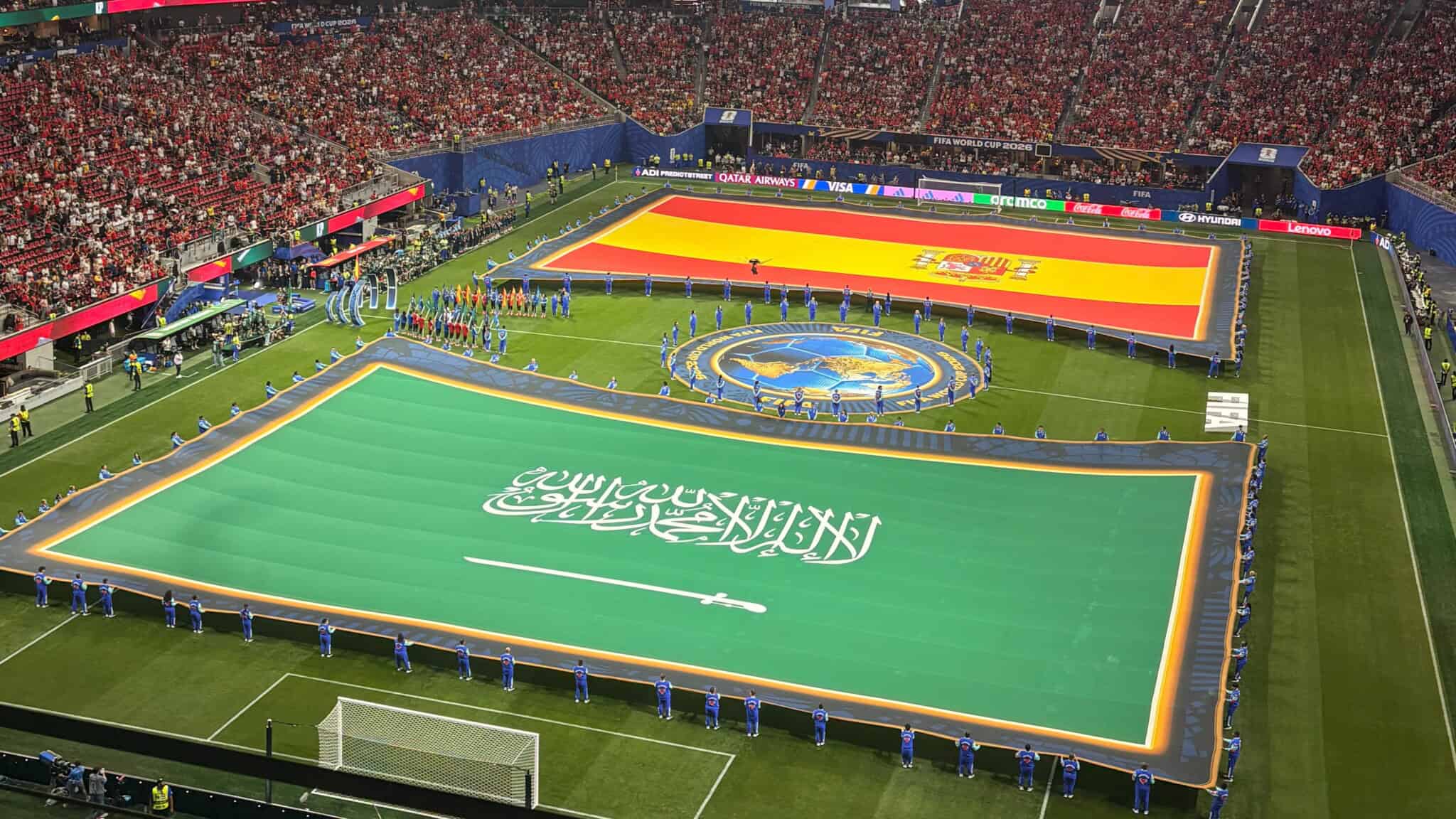 Large flags of Saudi Arabia and Spain are displayed on a soccer field, surrounded by officials and a stadium full of spectators before a match.
